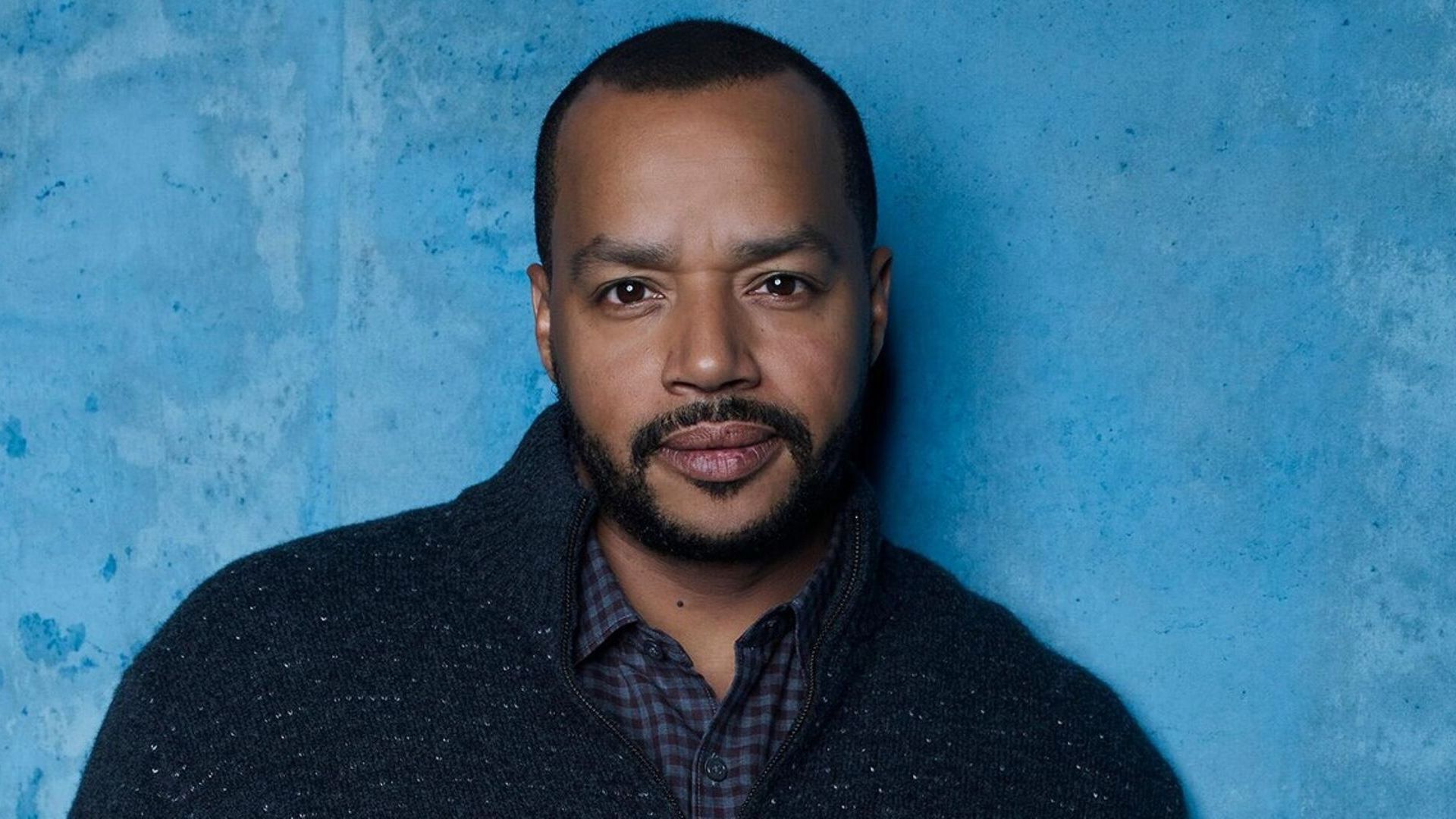 L'attore Donald Faison con un maglione blu su sfondo azzurro