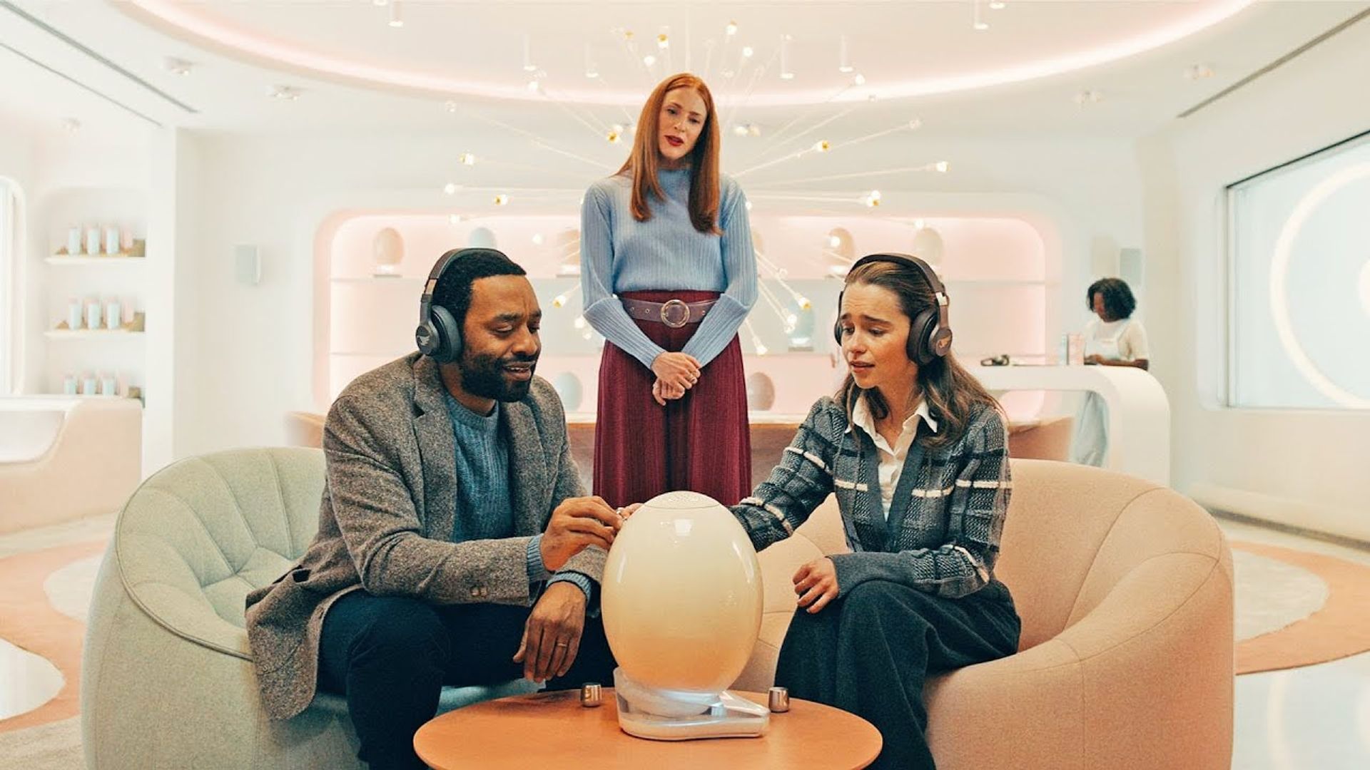 Emilia Clarke e Chiwetel Ejiofor seduti su delle sedie, ambientazione futuristica, scena da The Pod Generation