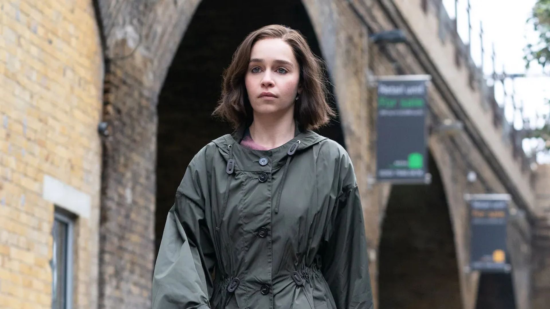 Emilia Clarke in impermeabile che cammina per strada, sguardo concentrato davanti a sé