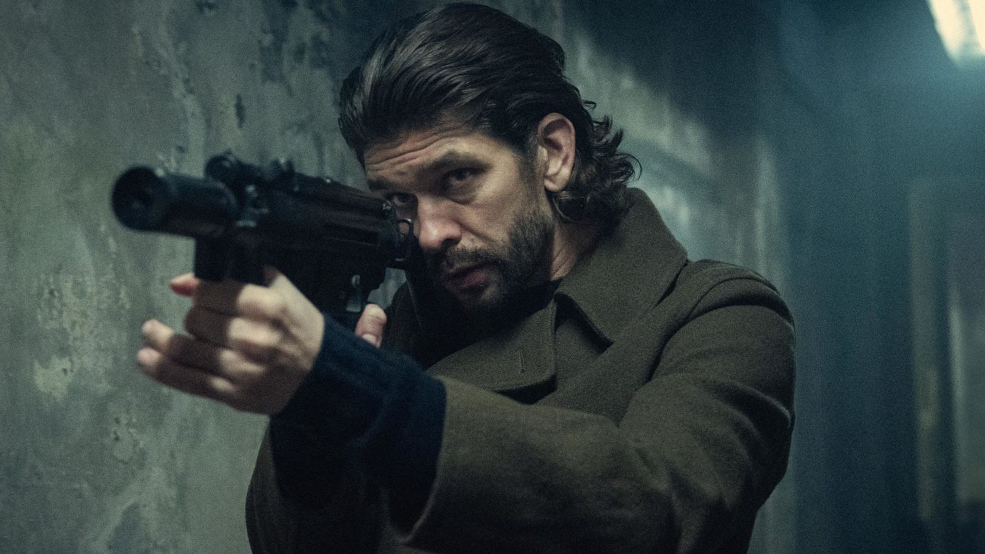 Ben Whishaw con capelli lunghi in cappotto che impugna una pistola