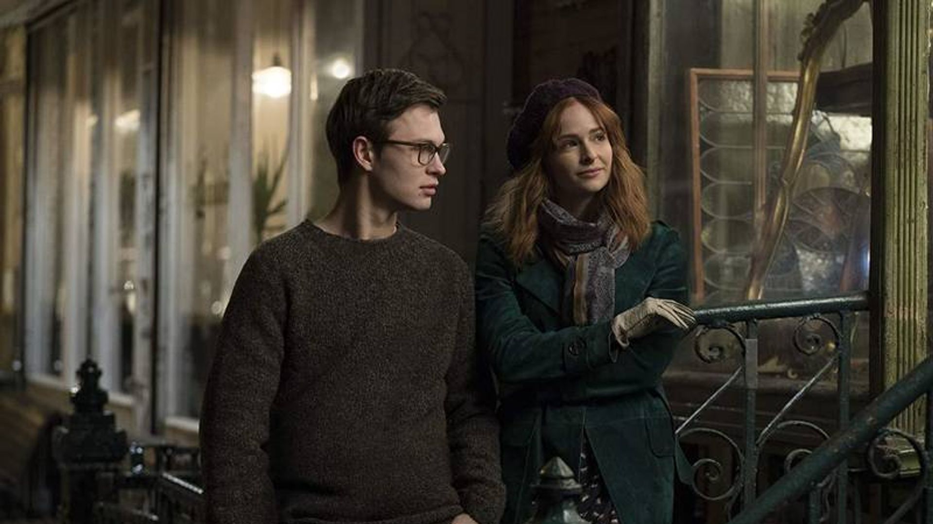 Un giovane uomo con gli occhiali e una donna con cappotto e basco che parlano in piedi vicino a una ringhiera in strada di sera