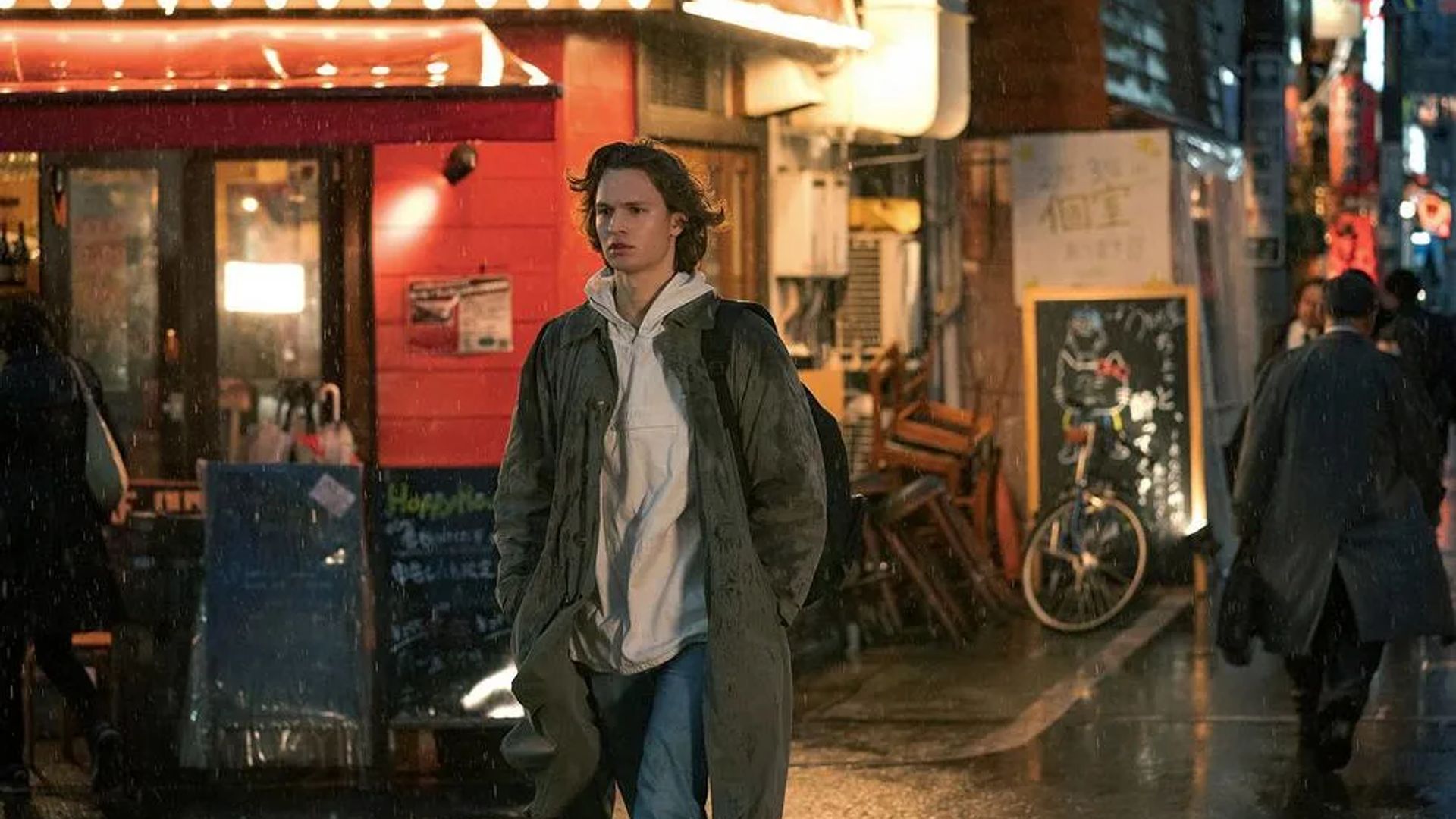 Ansel Elgort con cappotto e felpa con cappuccio cammina lungo una strada piovosa davanti a negozi e ristoranti illuminati