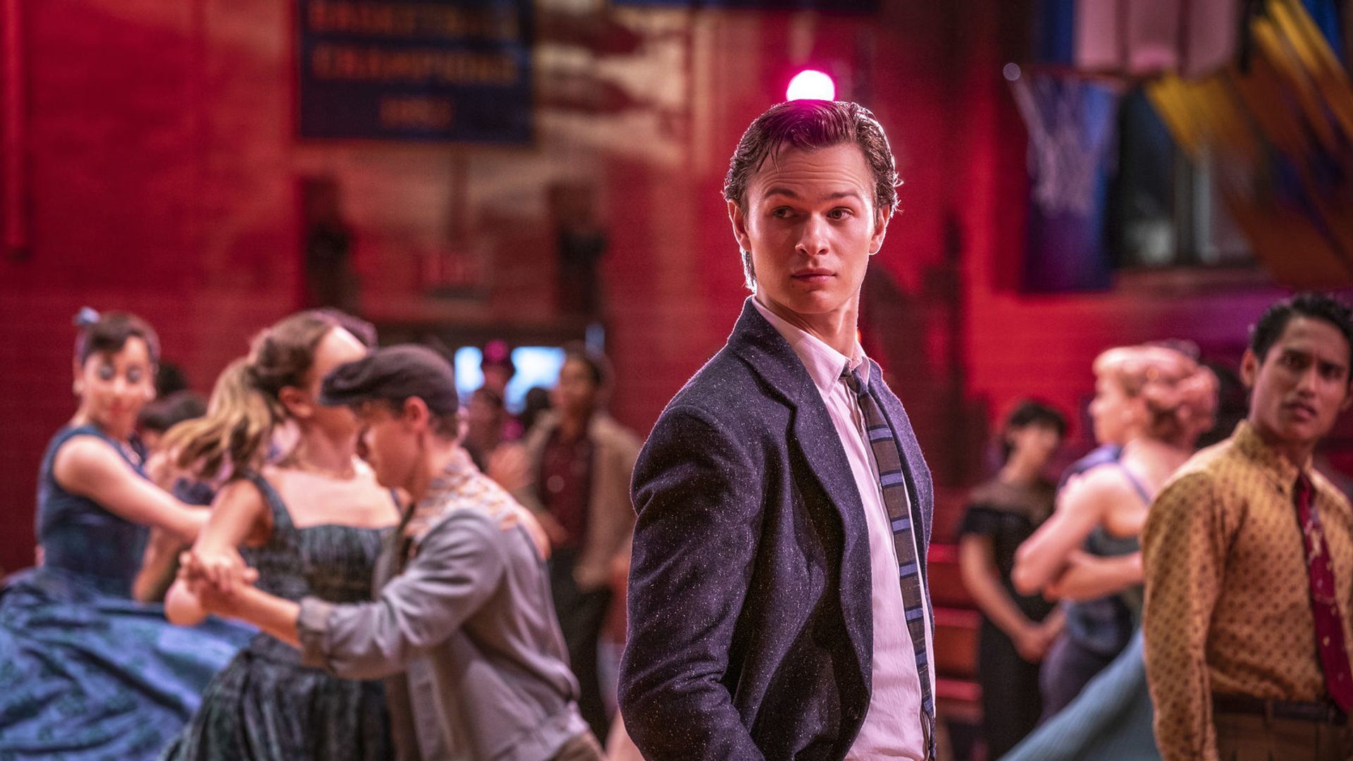 Ansel Elgort in giacca e cravatta in piedi a un ballo scolastico con coppie che ballano intorno a lui