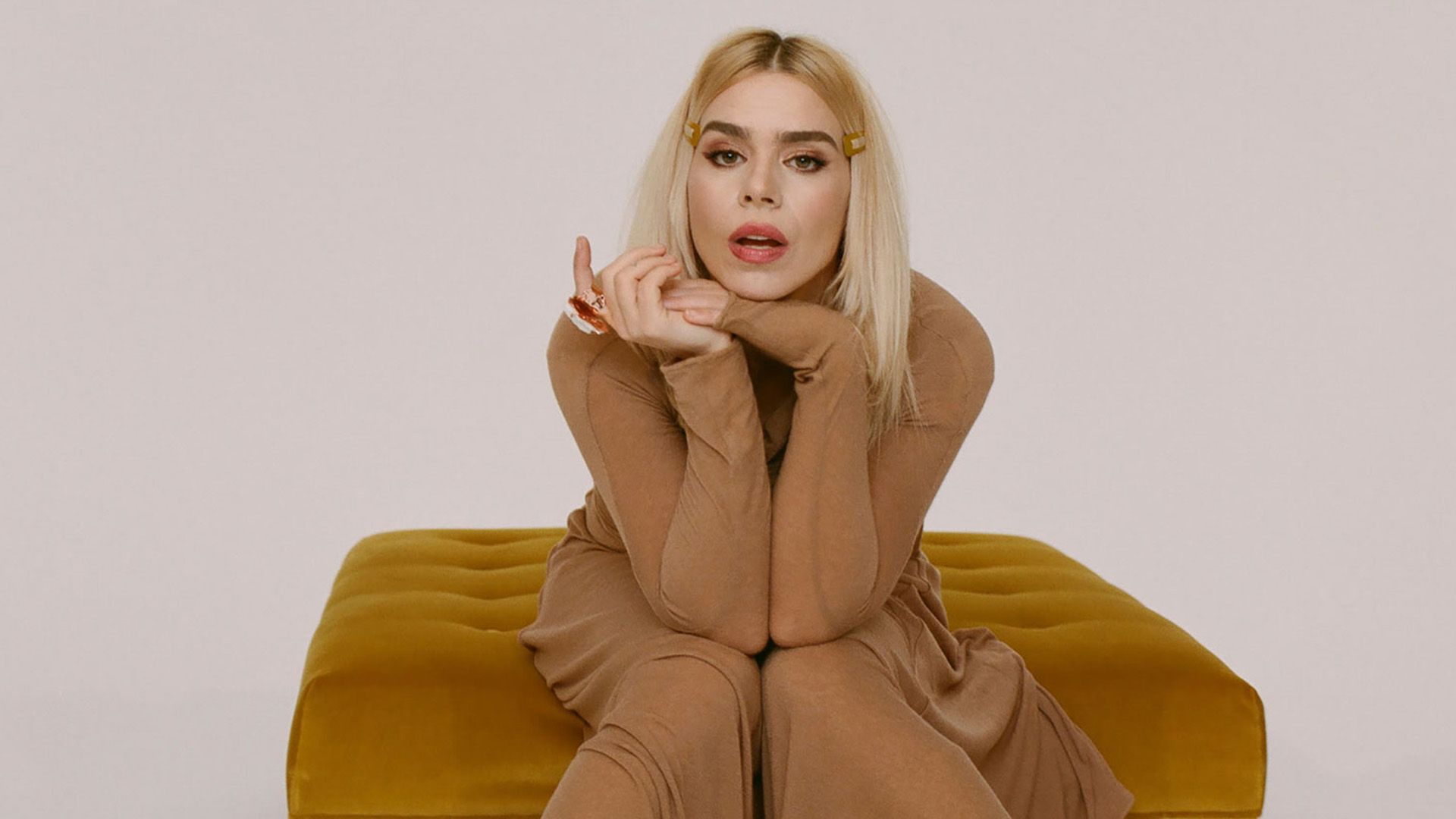 Billie Piper in un lungo abito beige seduta su una poltrona color zenzero