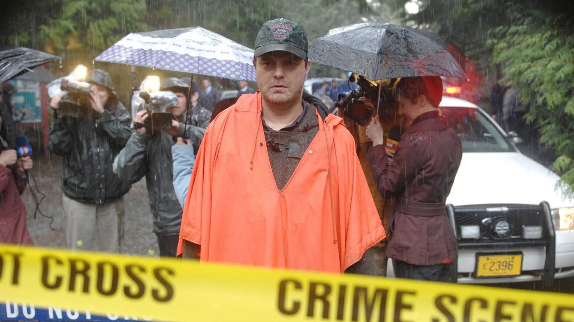 Rainn Wilson con un impermeabile arancione in piedi sotto la pioggia dietro il nastro giallo della polizia