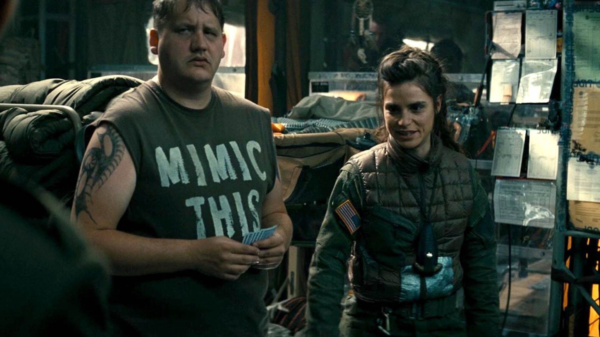 Charlotte Riley in uniforme kaki accanto a un uomo grande in una canottiera nera