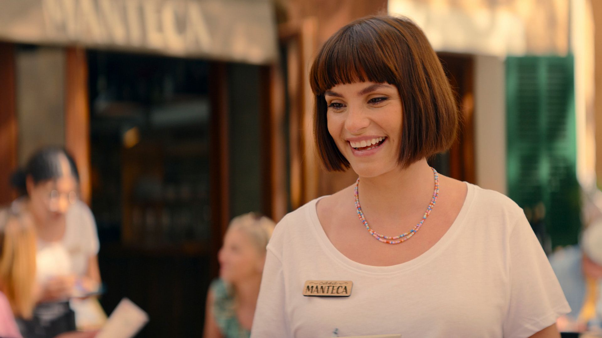 Charlotte Riley con taglio bob che indossa una t-shirt bianca, un badge di legno sul petto e una collana di perline colorate