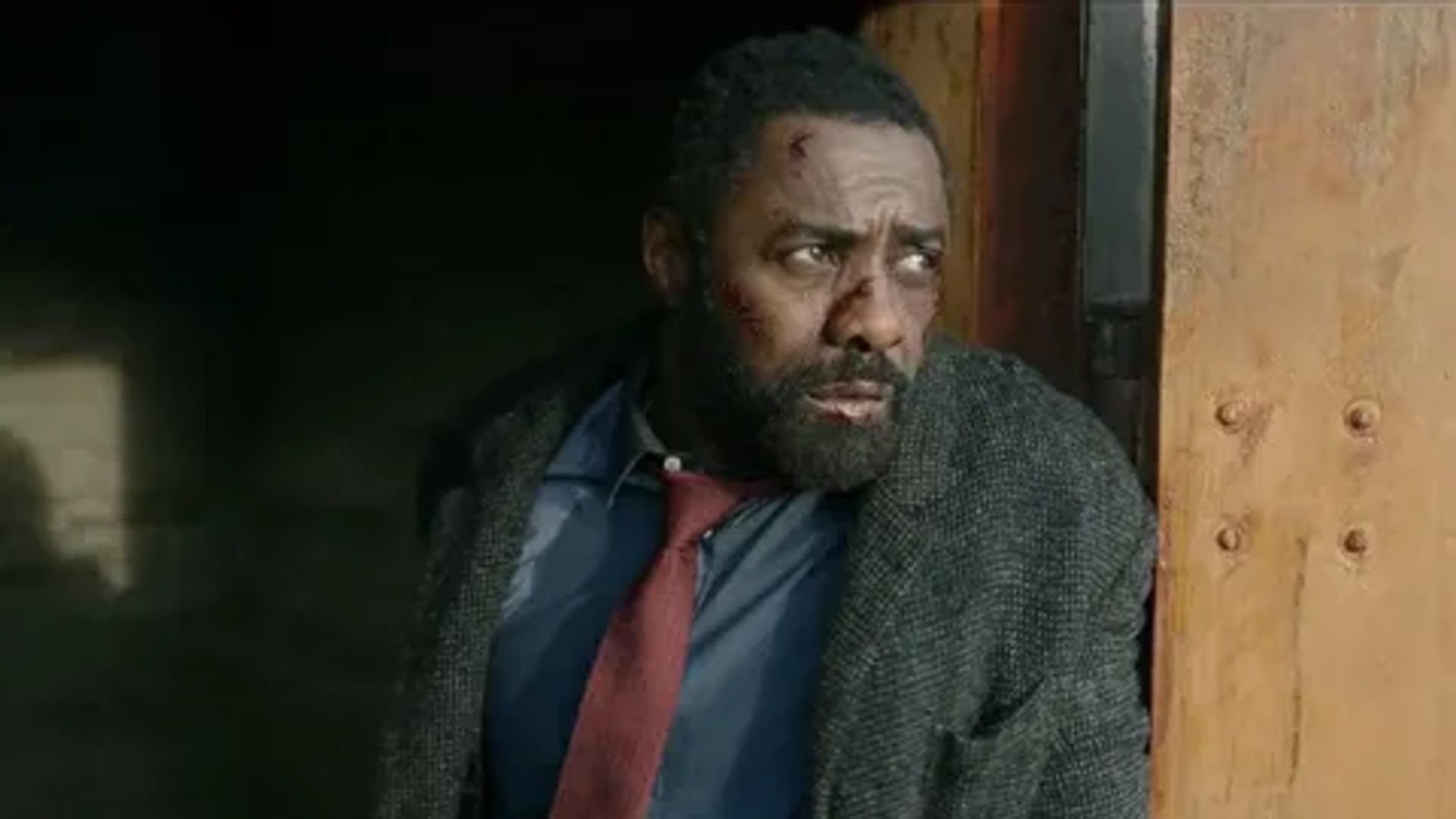 Idris Elba con cappotto e cravatta rossa che guarda di lato, fotogramma del film
