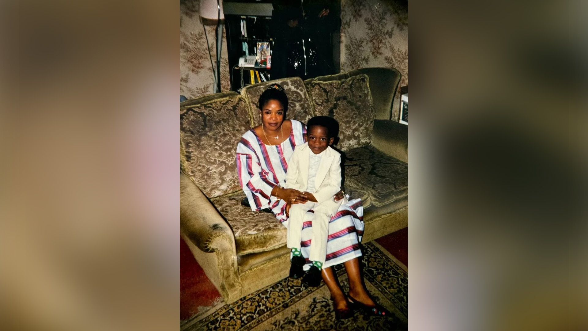 Il piccolo Damson Idris in abito bianco siede sulle ginocchia di sua madre seduta sul divano
