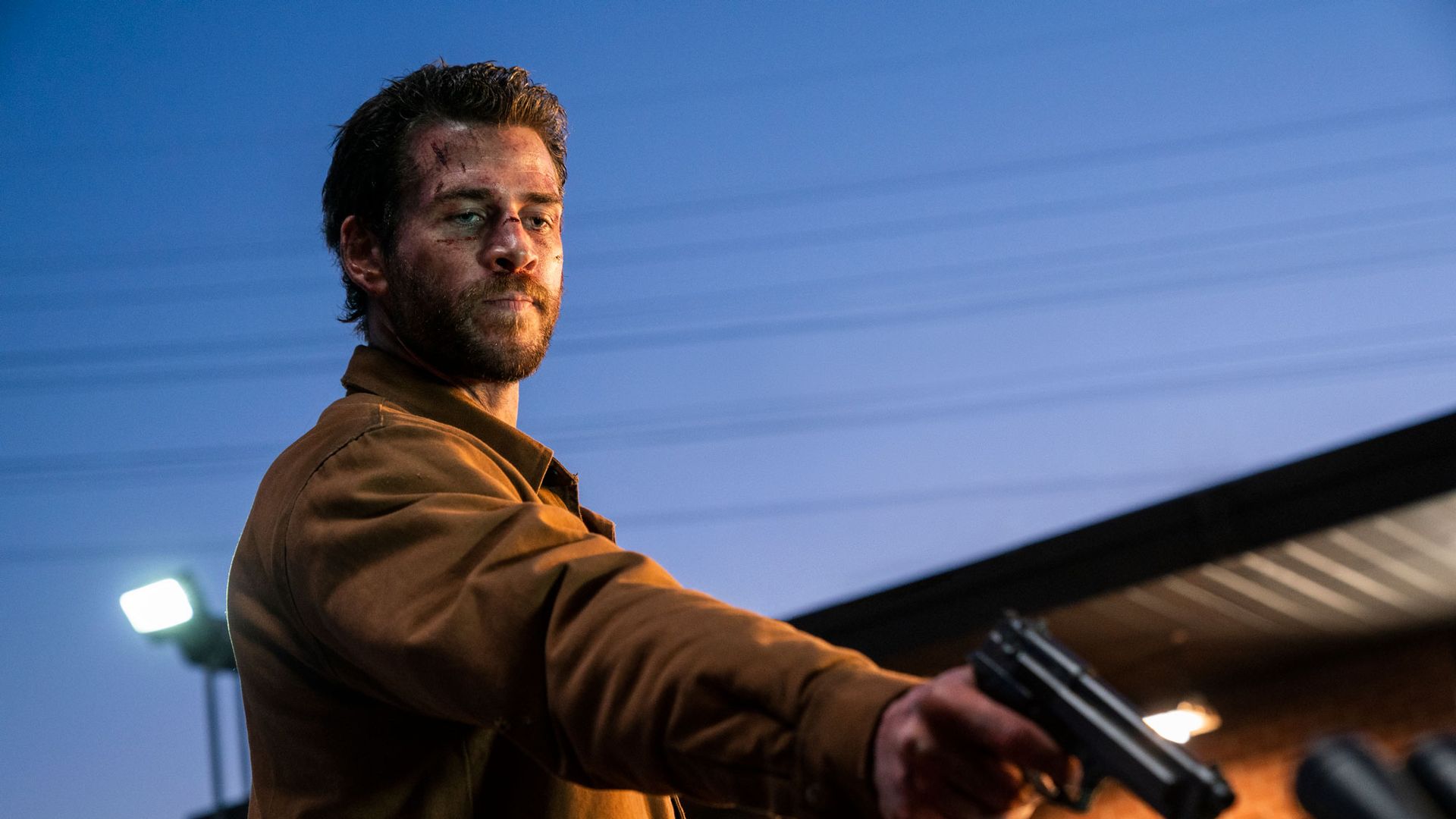 Liam Hemsworth con una giacca marrone che impugna una pistola