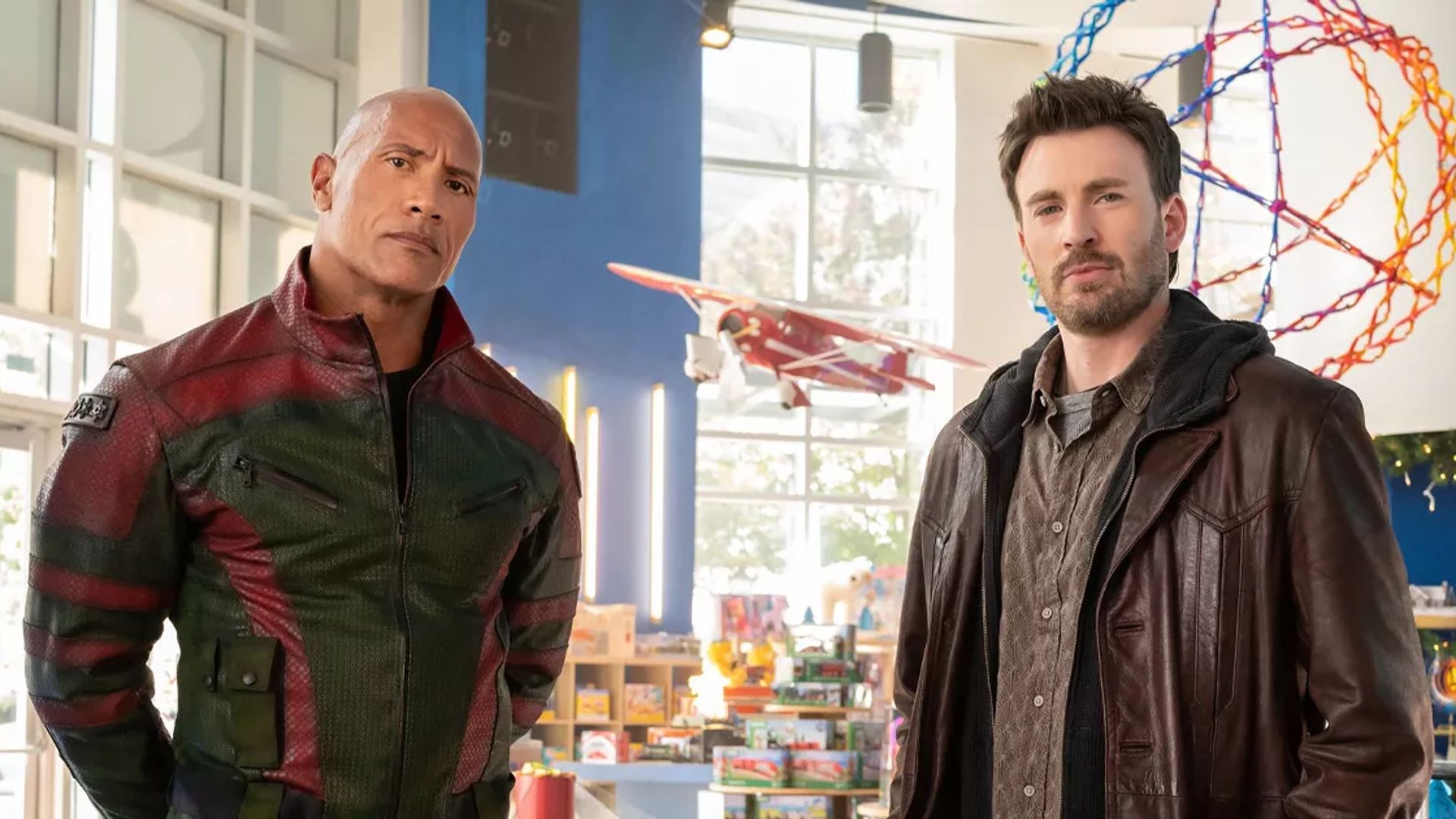 Chris Evans e Dwayne Johnson con giacche in una stanza illuminata con giocattoli e un modellino di aereo sullo sfondo
