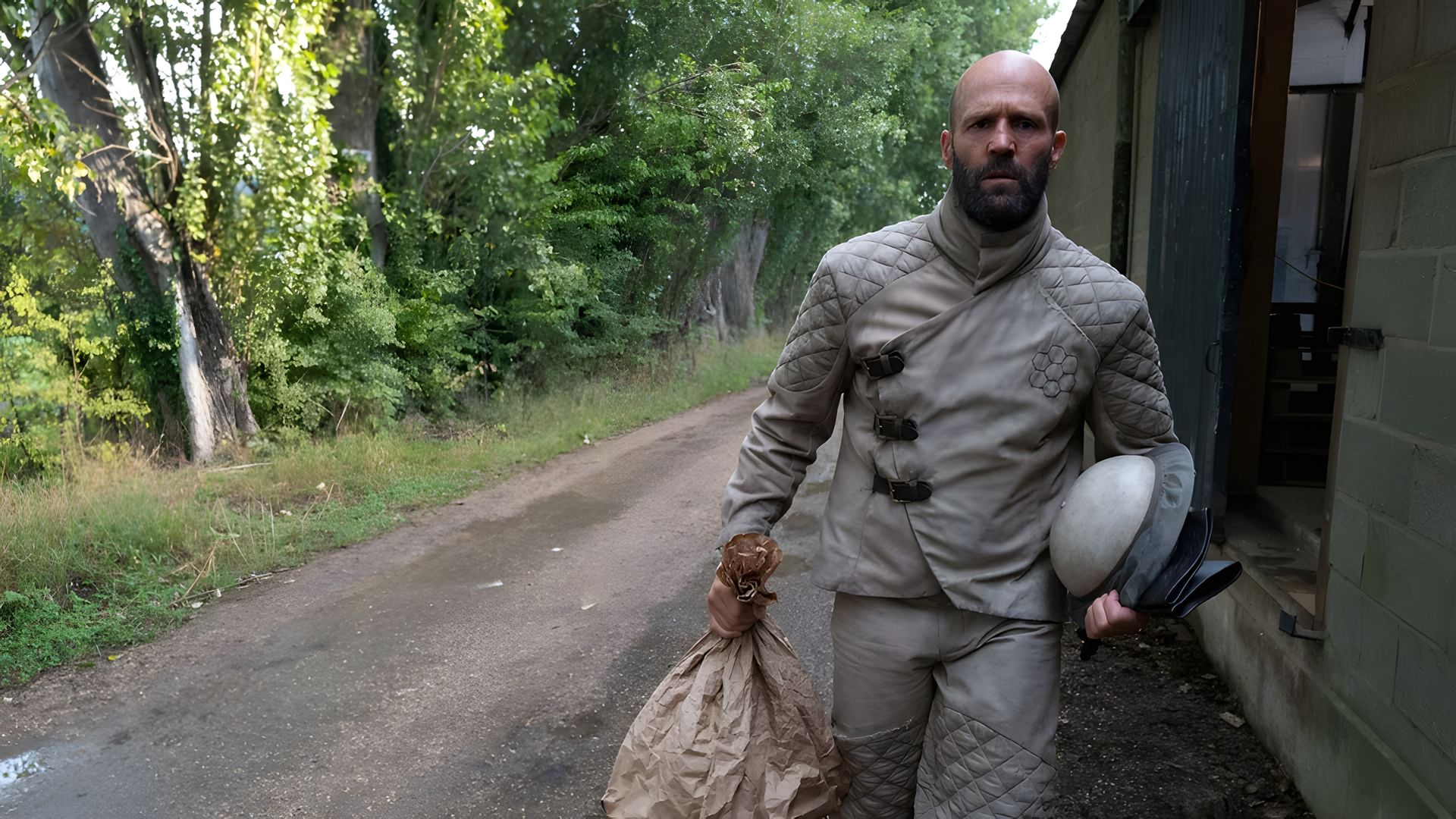 Jason Statham in tuta protettiva grigia cammina lungo una strada portando una borsa e un casco con alberi sullo sfondo