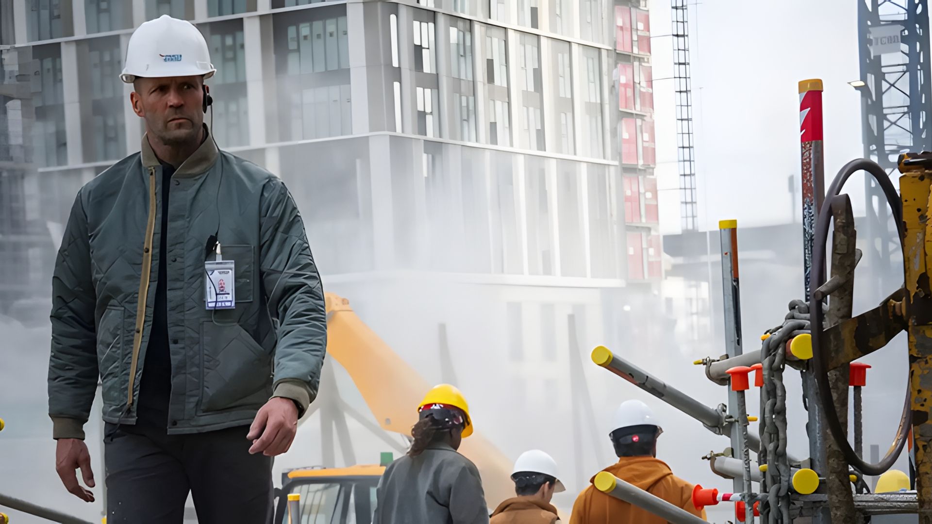 Jason Statham con un casco e una giacca cammina attraverso un cantiere edile con operai e macchinari visibili sullo sfondo nebbioso