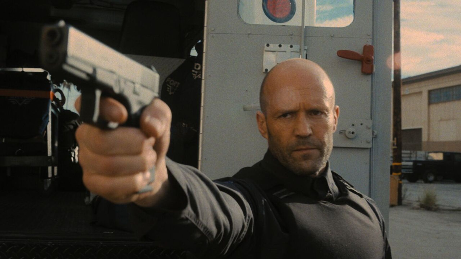 Jason Statham in equipaggiamento tattico nero punta una pistola mentre si trova accanto a un furgone blindato su uno sfondo industriale