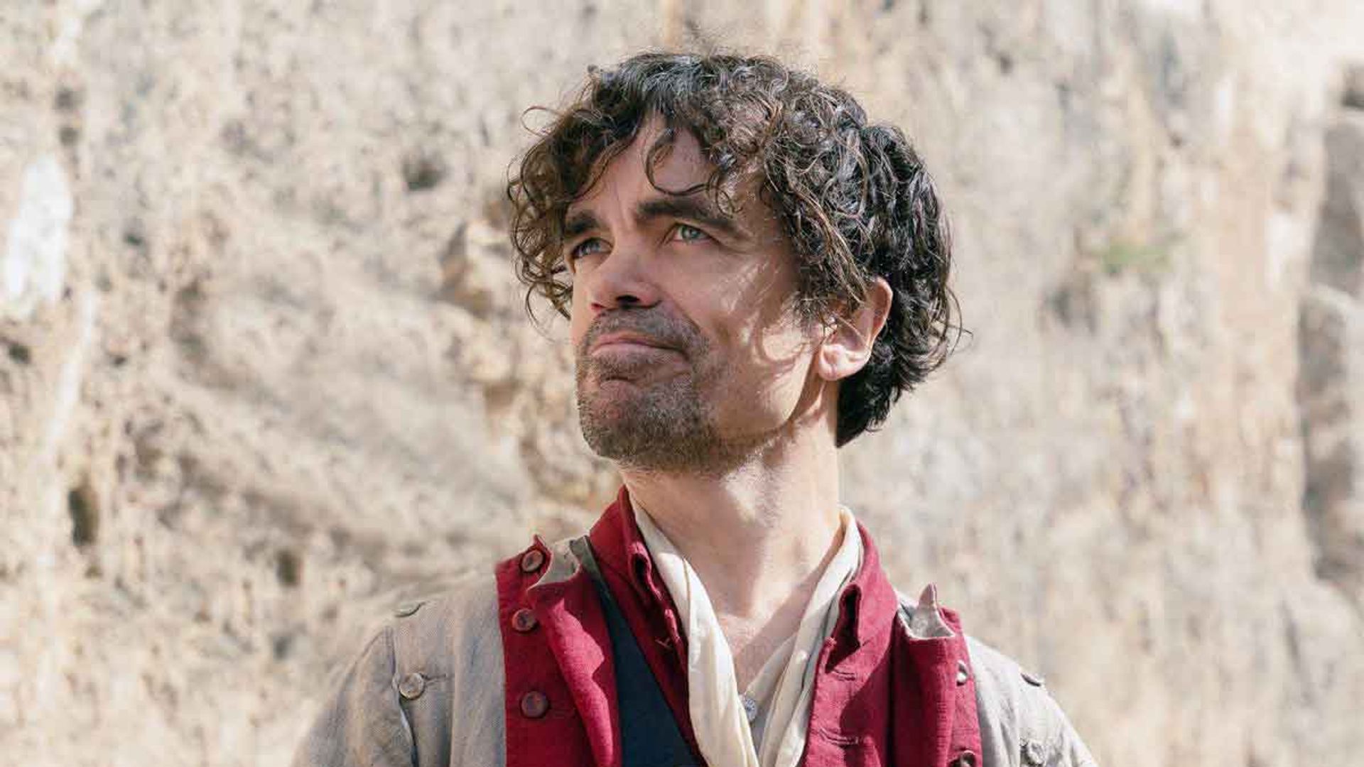 Peter Dinklage in costume d'epoca con un farsetto rosso tiene una lettera e guarda in alto con un leggero sorriso