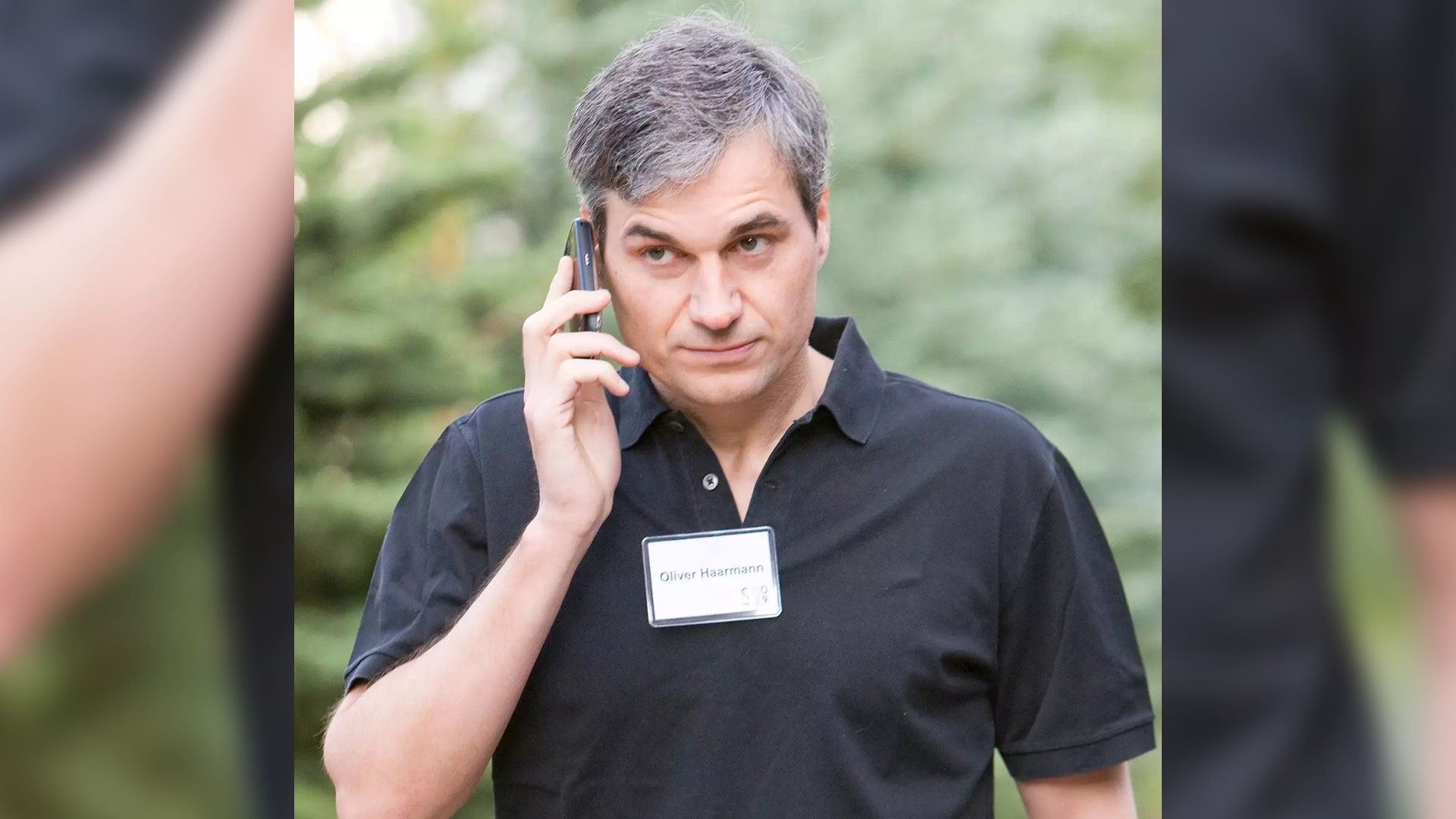 Un uomo in camicia nera con un distintivo che parla al telefono sullo sfondo di alberi verdi, con espressione concentrata