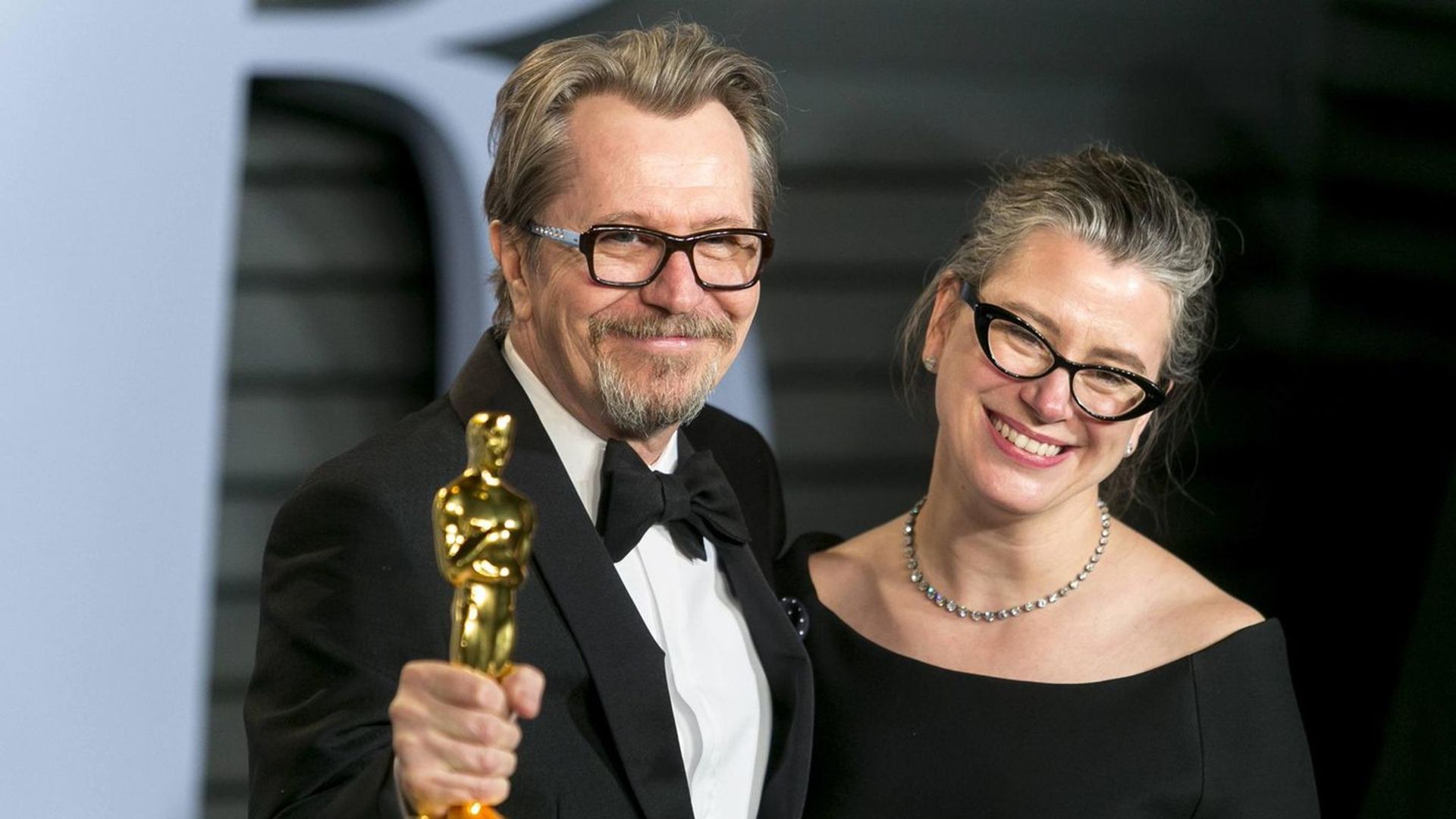 Gary Oldman in smoking tiene in mano una statuetta dell'Oscar e sorride accanto a Gisele Schmidt in abito nero e occhiali