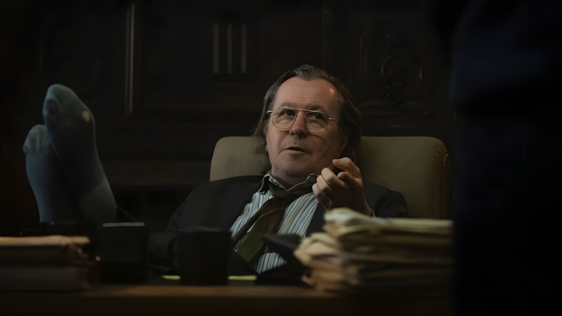 Gary Oldman con occhiali e abito seduto alla scrivania in un ufficio con i piedi sul tavolo, con un'espressione pensierosa