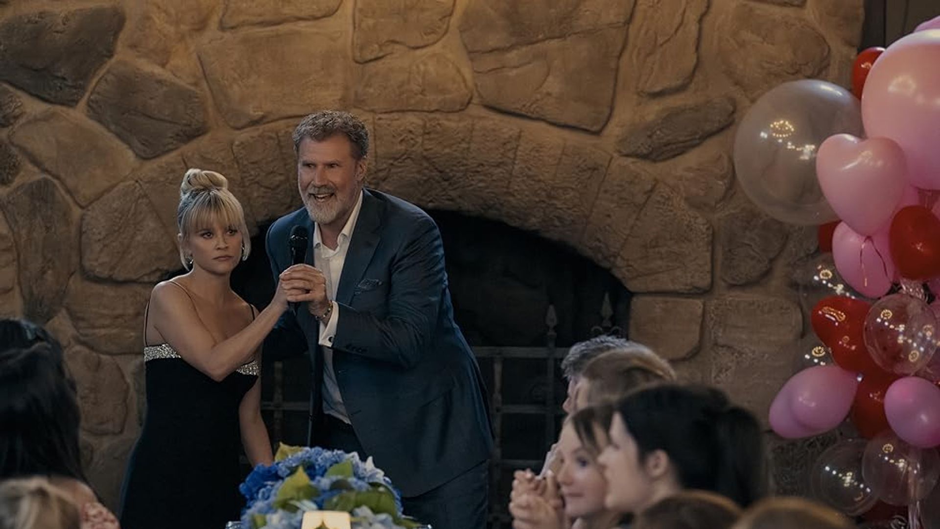 Will Ferrell in giacca e cravatta con un microfono tiene per mano Reese Witherspoon in abito nero sullo sfondo di un camino, tavola festiva e palloncini