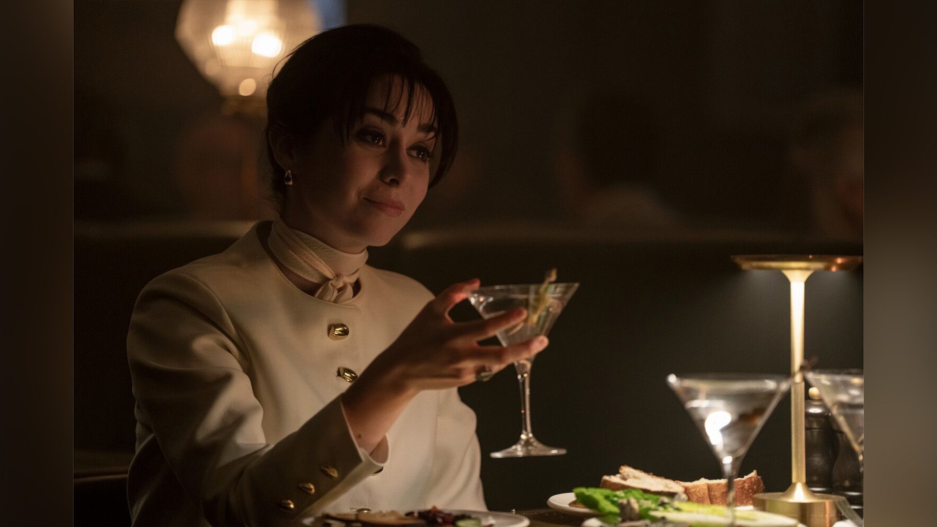 Cristin Milioti in un abito bianco che tiene un bicchiere in una scena della serie "The Penguin"