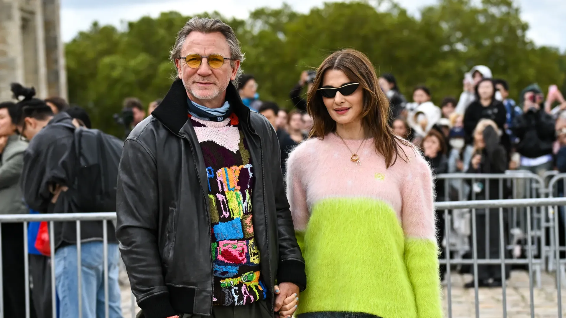 Rachel Weisz in un maglione bianco e verde e Daniel Craig in una giacca di pelle nera e maglione colorato che si tengono per mano