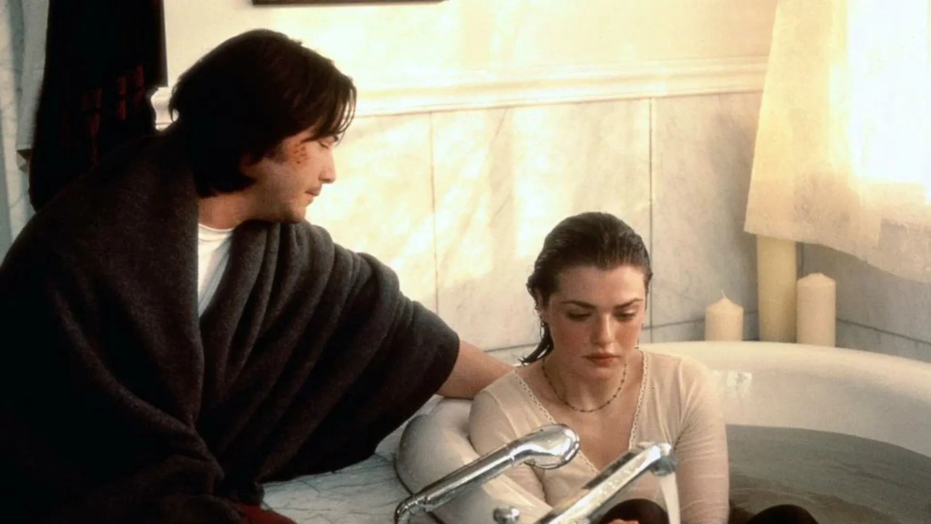 La giovane Rachel Weisz è seduta nella vasca da bagno vestita, accanto a lei siede il giovane Keanu Reeves