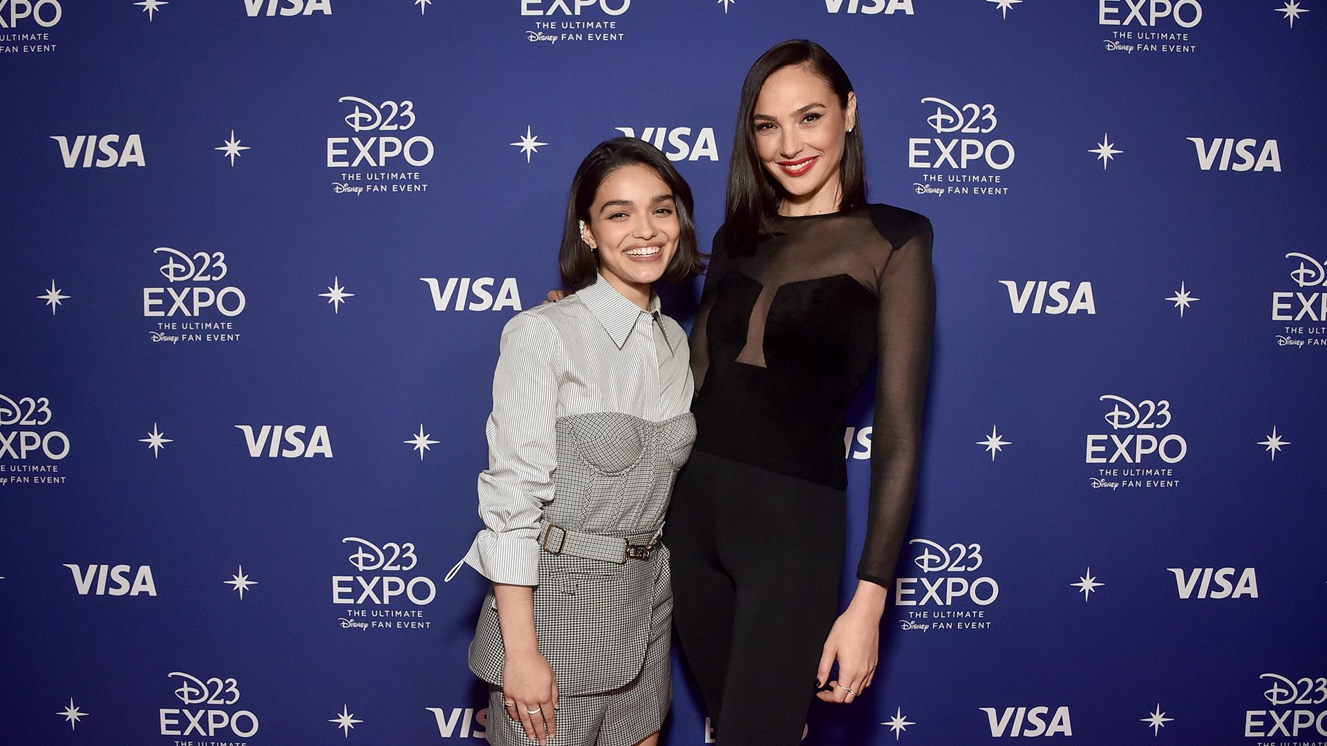 Rachel Zegler in abiti chiari e Gal Gadot in nero contro uno sfondo blu ridono