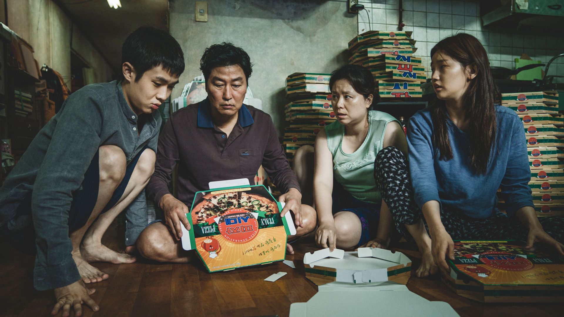 Una famiglia sudcoreana di quattro persone seduta per terra circondata da scatole di pizza in una scena del film "Parasite"