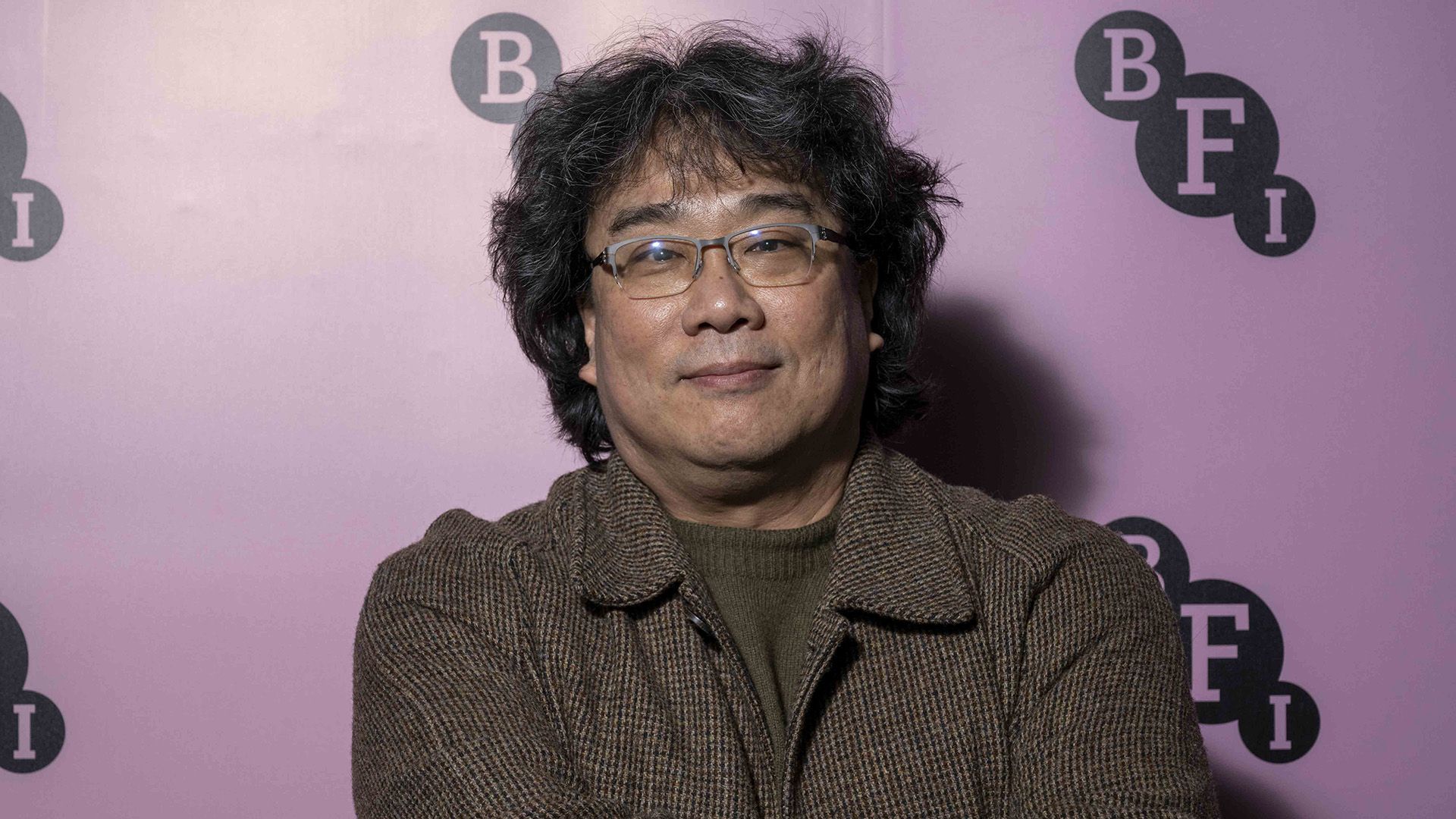 Director Bong Joon Ho in a brown jacket and glasses against a lilac background
