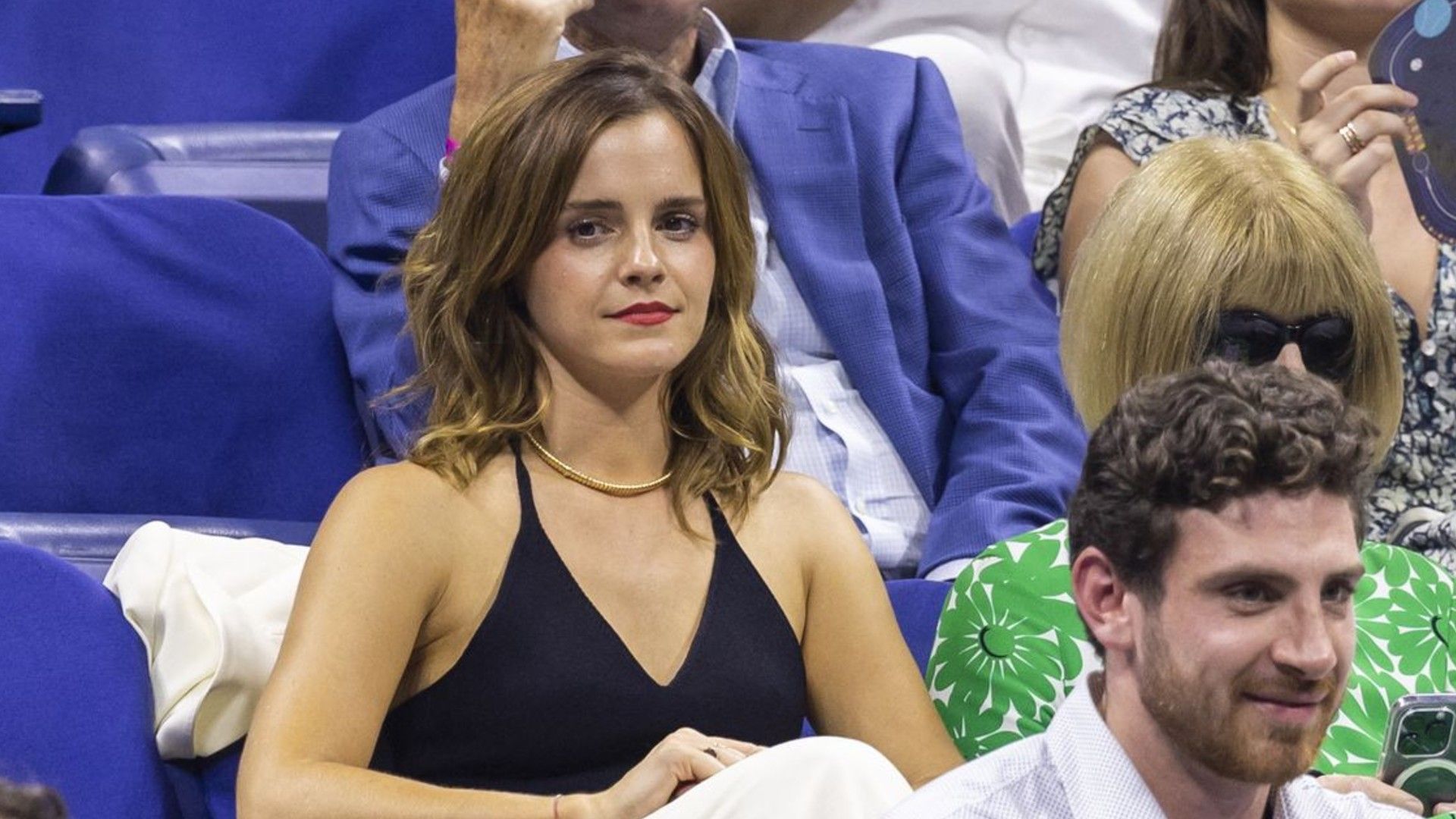 Emma Watson sugli spalti di un evento sportivo, guarda avanti, seduta con un top nero e una collana d'oro