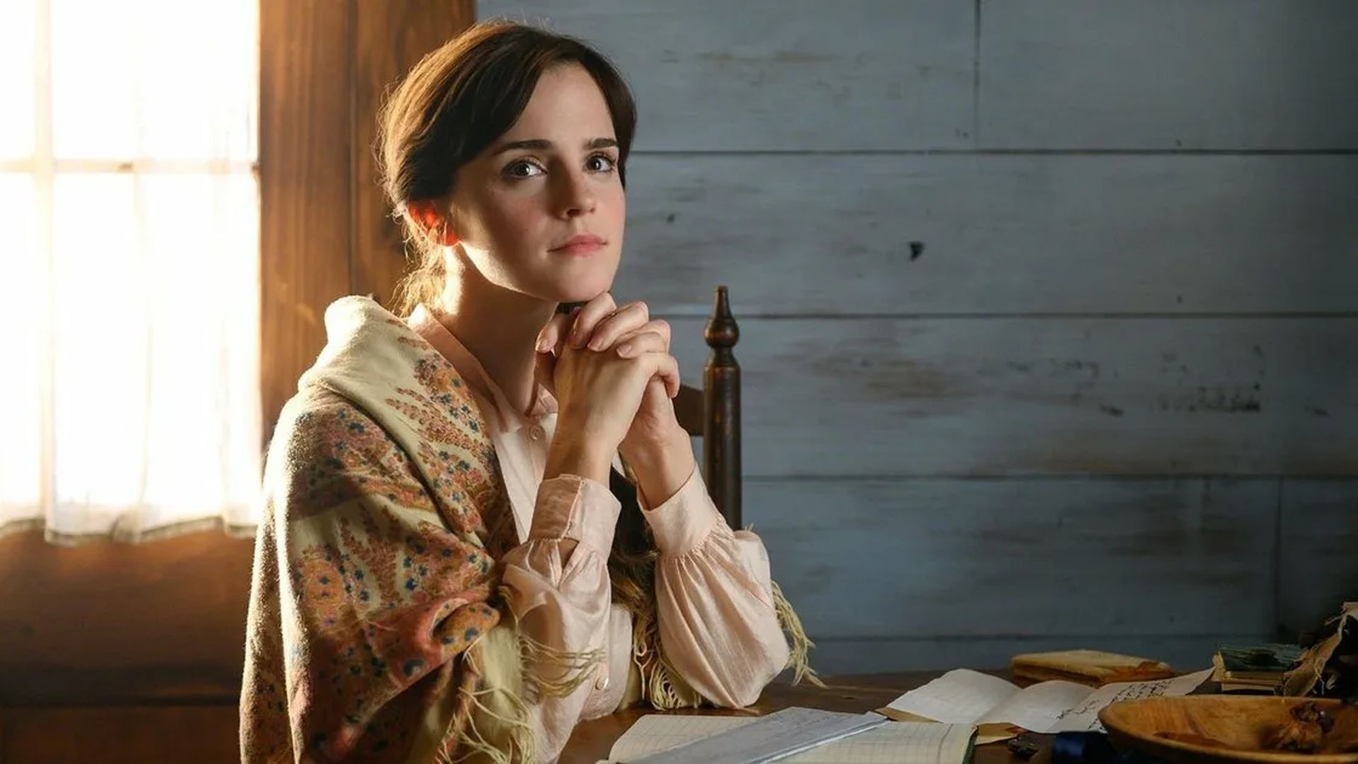 Emma Watson seduta al tavolo con un'espressione pensierosa sul volto, mani giunte