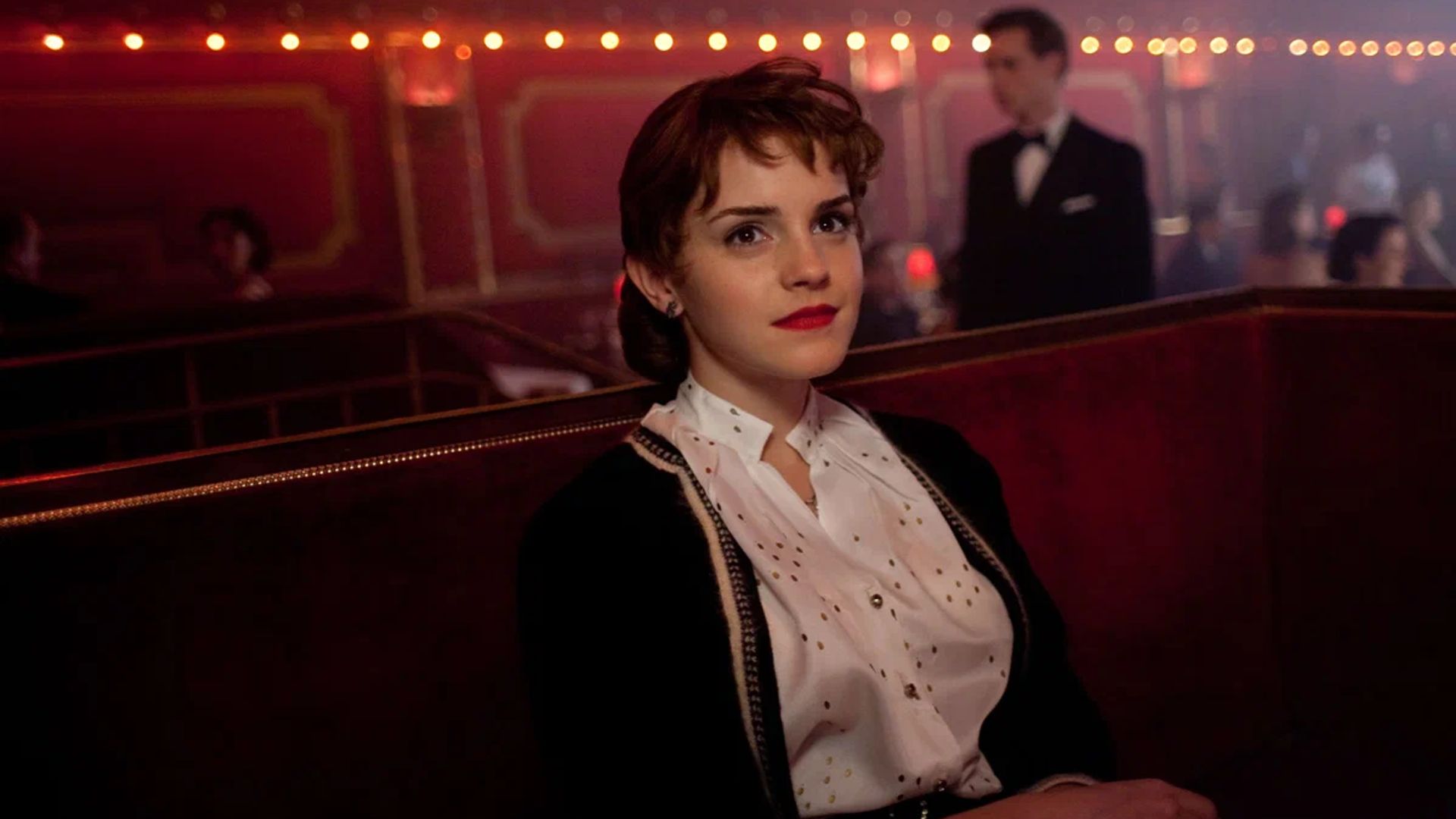 Emma Watson con un'acconciatura e rossetto rosso seduta in un club scuro