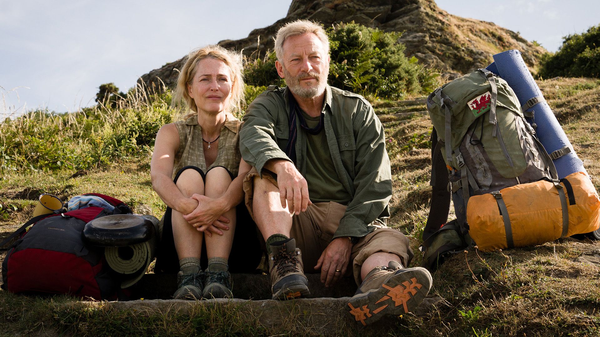 Gillian Anderson e Jason Isaacs sono seduti su una collina, i loro zaini da trekking sono accanto a loro