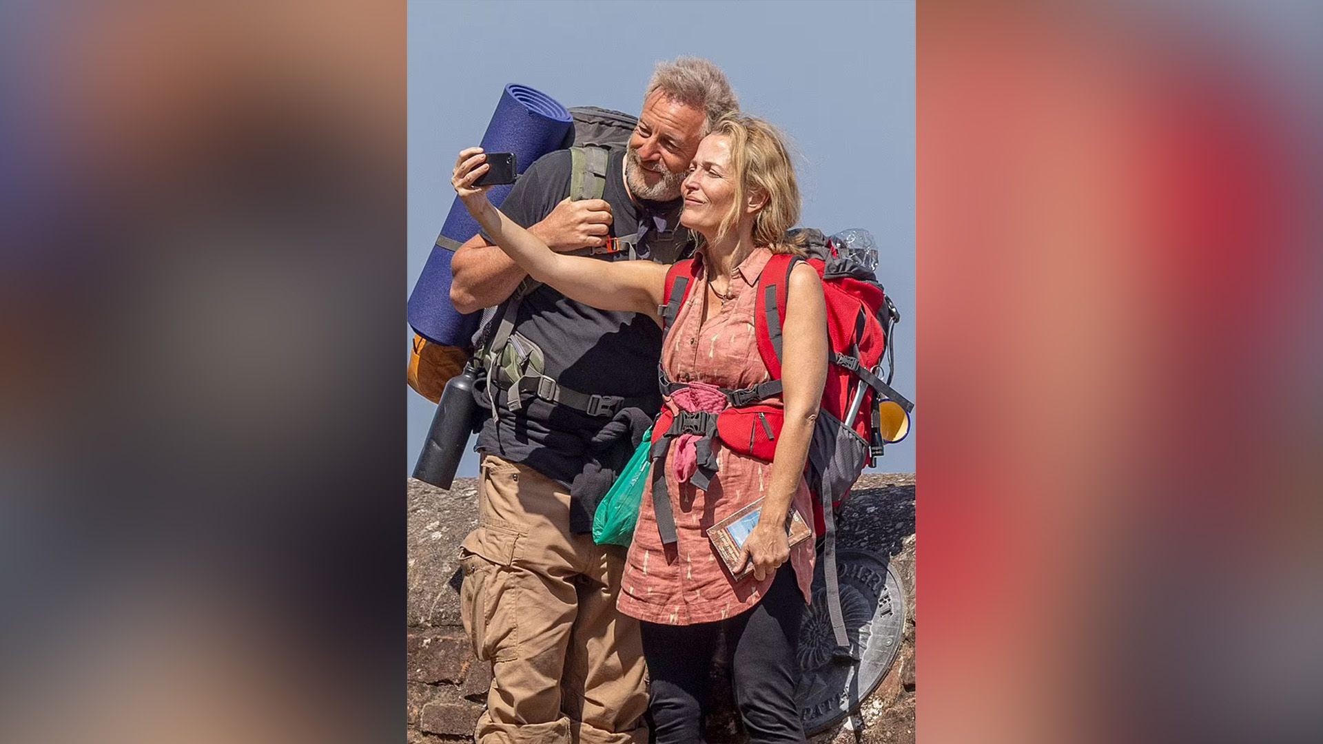 Jason Isaacs e Gillian Anderson con zaini sulle spalle stanno facendo un selfie al telefono