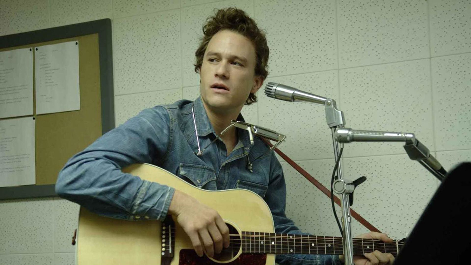 Heath Ledger con una chitarra e un'armonica davanti a un microfono nel film "I'm Not There"