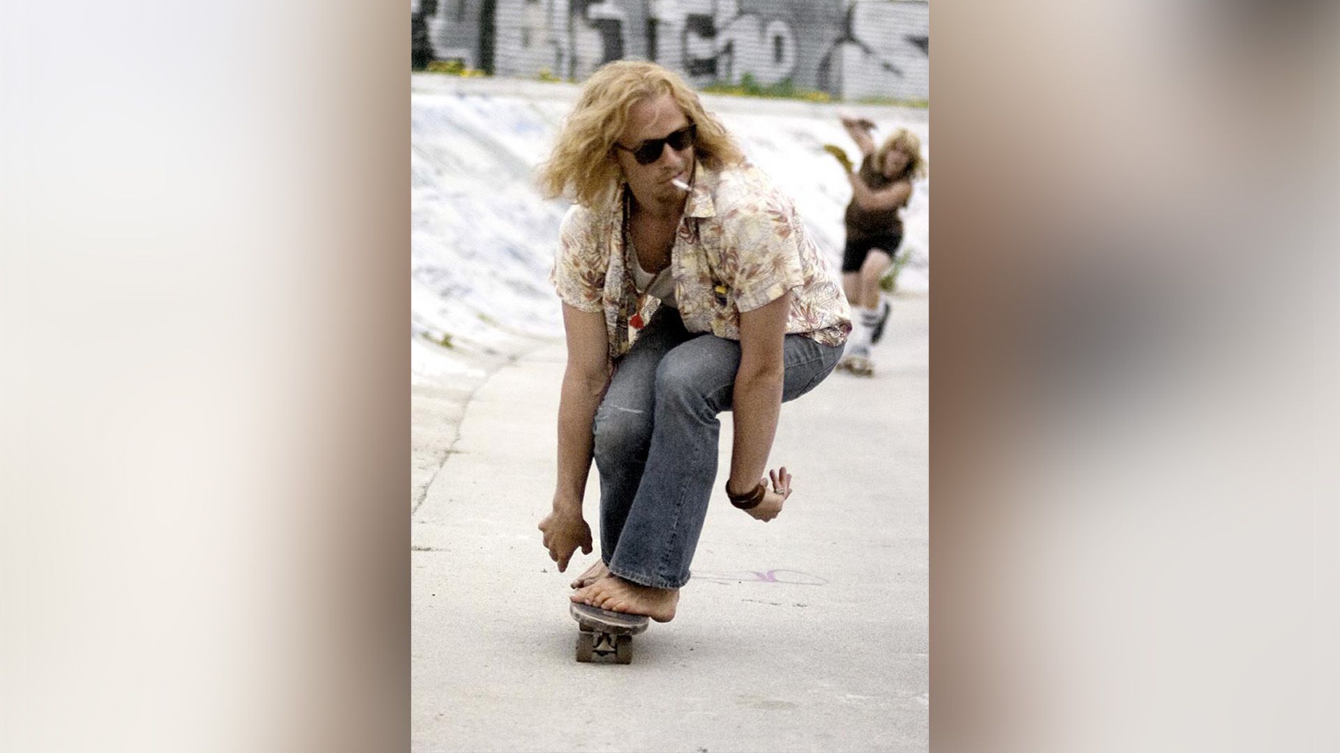 Heath Ledger con i capelli lunghi e occhiali scuri che va in skateboard a piedi nudi