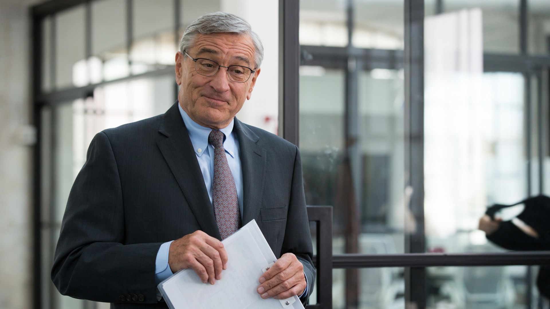 Robert De Niro in giacca, cravatta e occhiali tiene in mano una cartella per documenti