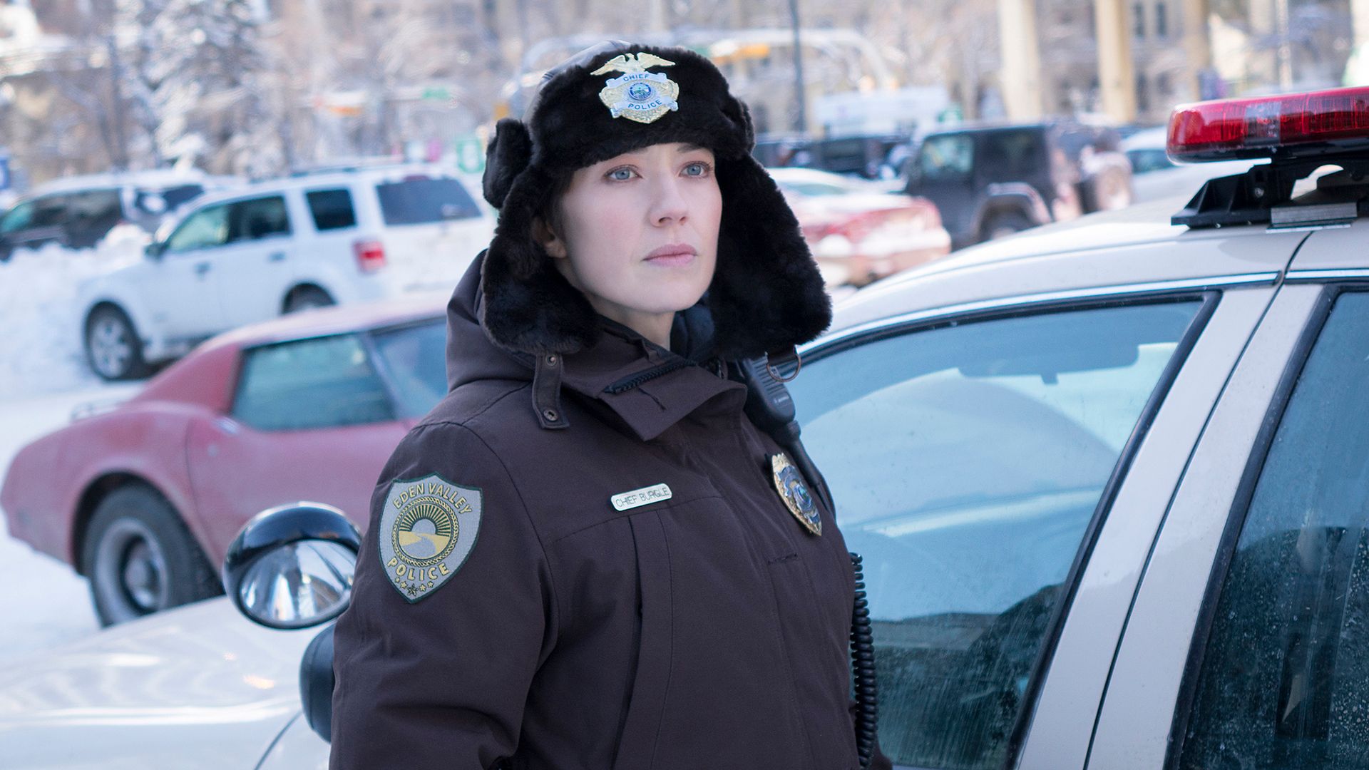 Carrie Coon in una divisa invernale da poliziotto accanto a un'auto della polizia