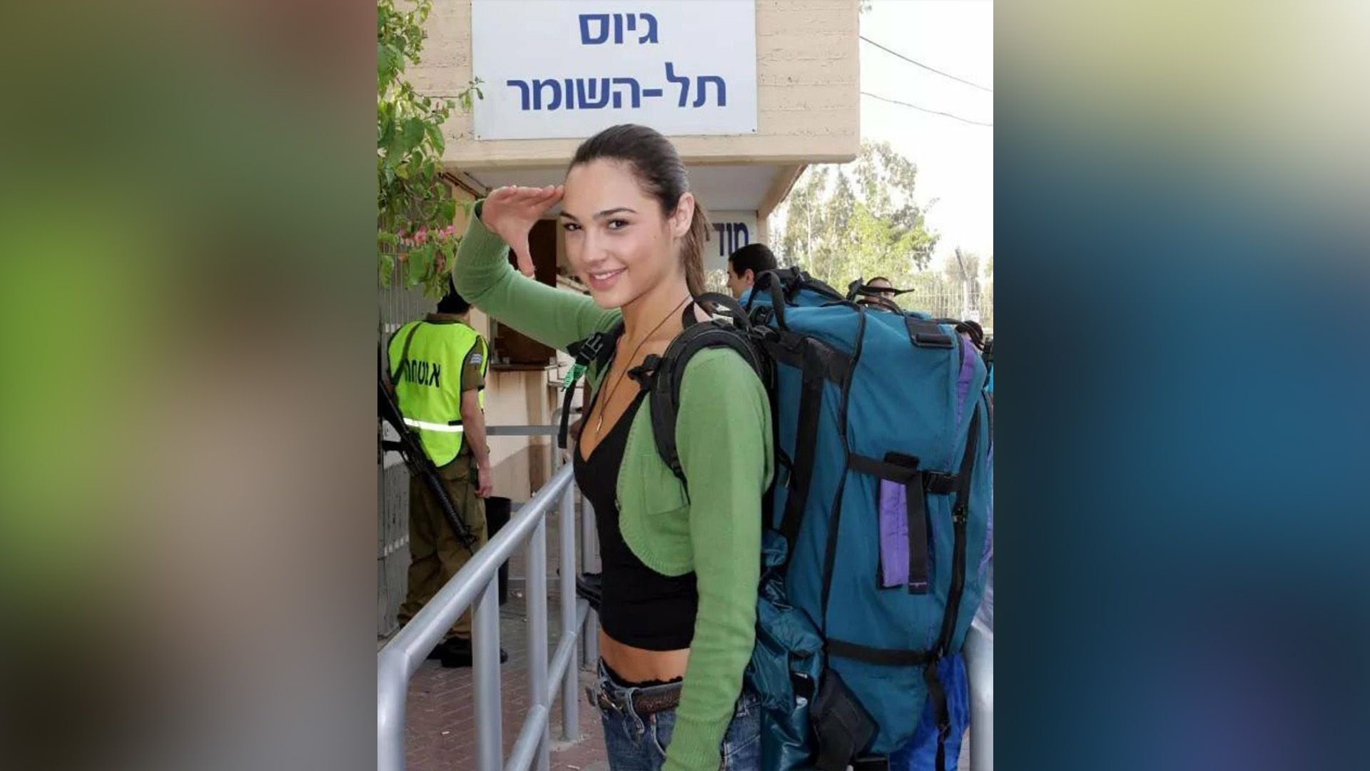 Giovane Gal Gadot in un maglione verde e con un grande zaino blu sulla schiena che saluta con la mano destra