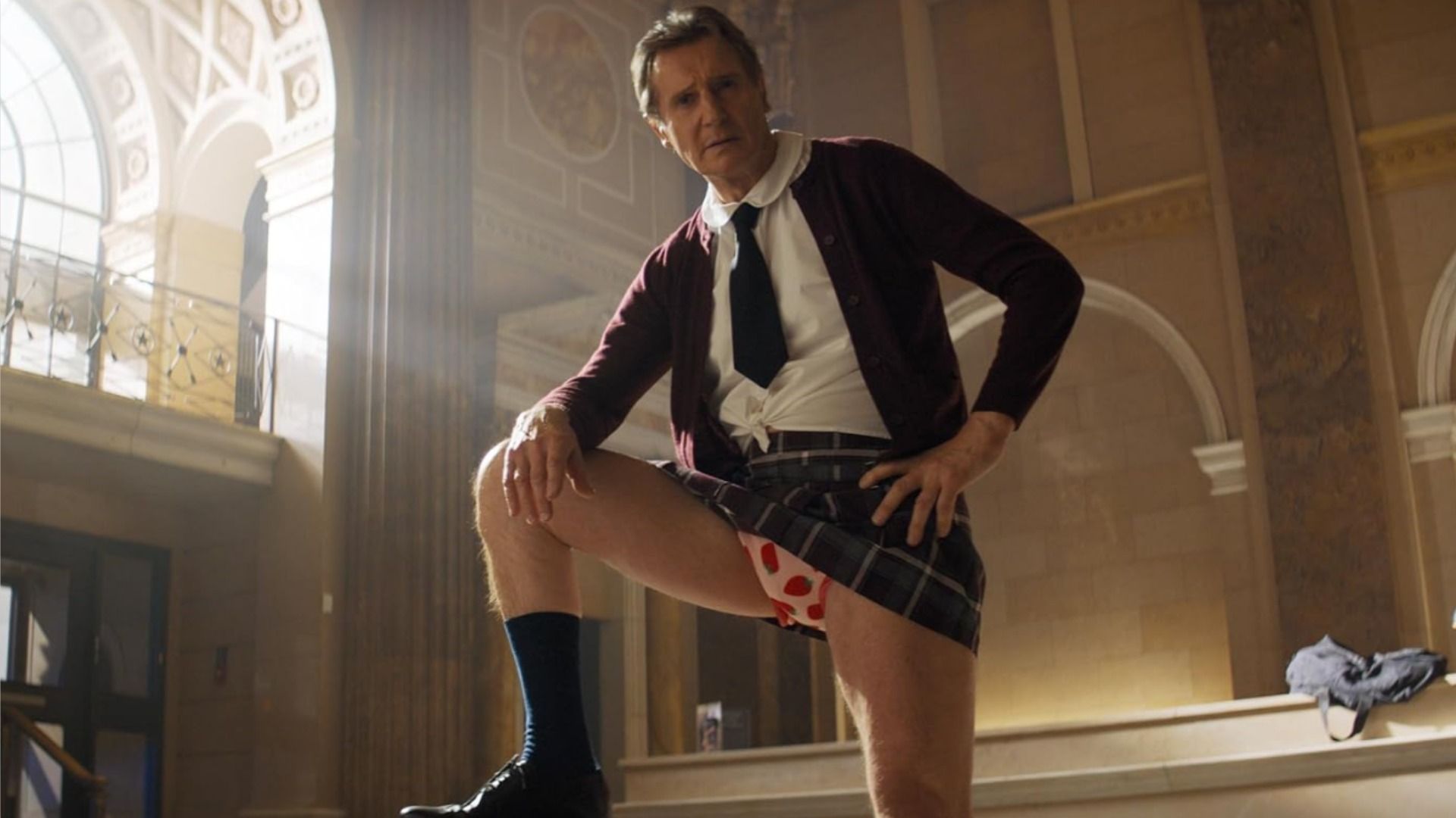 L'attore Liam Neeson in uniforme scolastica posa sullo sfondo di una banca
