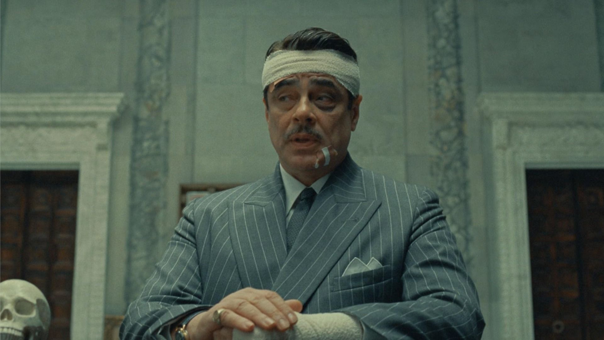 L'attore Benicio del Toro in una giacca grigia e con la testa fasciata seduto a un tavolo contro un muro