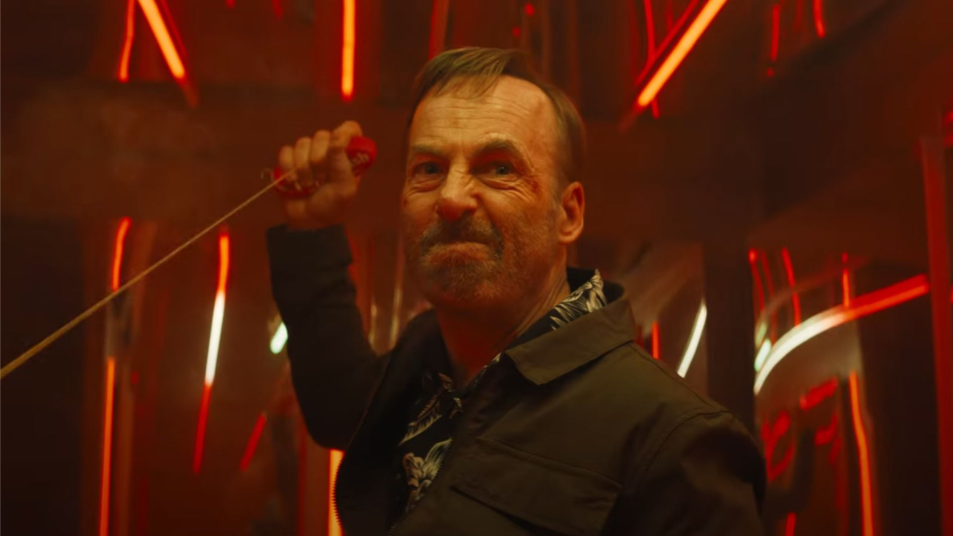 L'attore Bob Odenkirk in giacca nera tiene una corda nelle mani e guarda rabbioso nella direzione della luce rossa