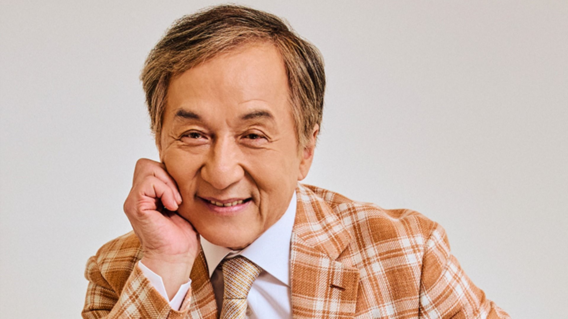 L'attore Jackie Chan in una giacca marrone a quadri siede, appoggiato sulla mano, e sorride alla fotocamera su uno sfondo bianco
