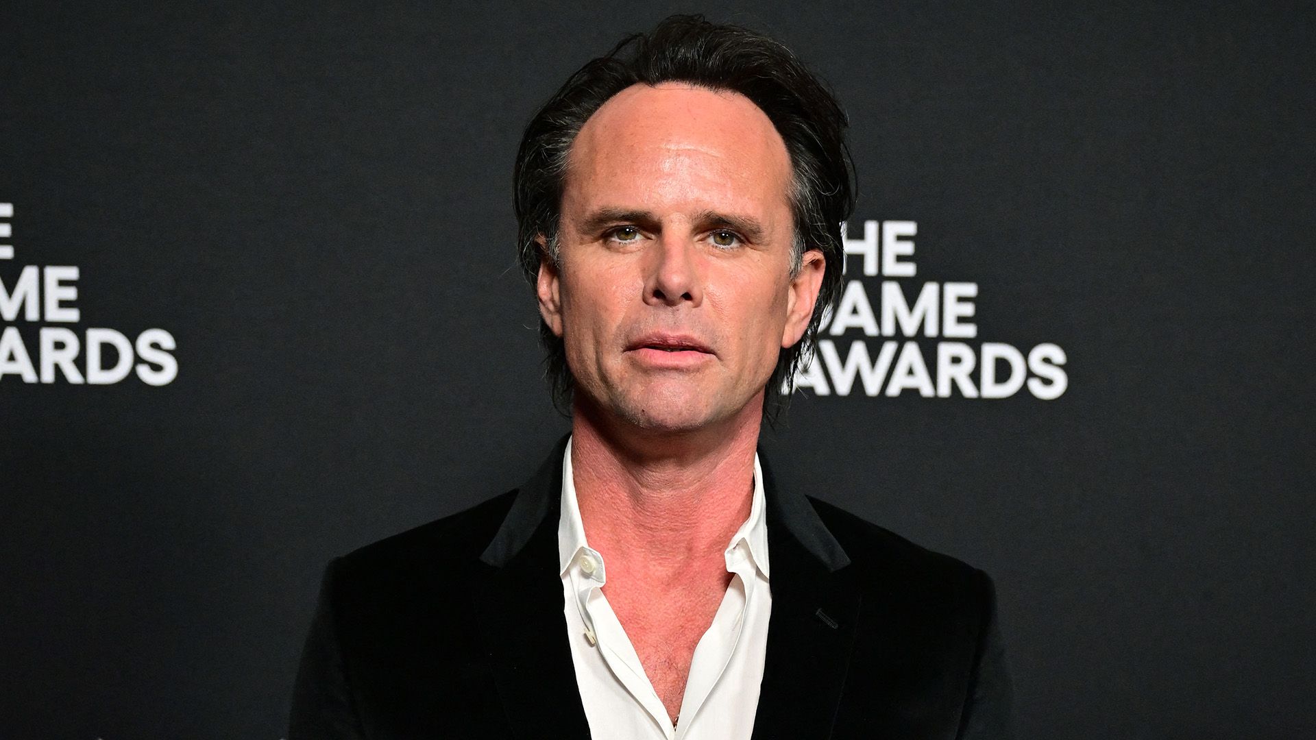 L'attore Walton Goggins in una giacca nera e camicia bianca con colletto sbottonato su sfondo nero