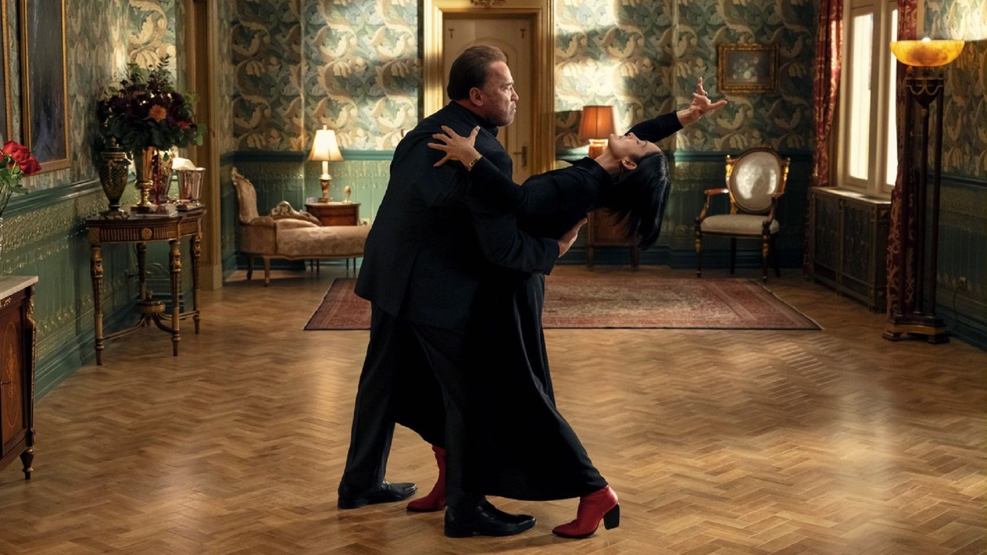 Arnold Schwarzenegger e Carrie-Anne Moss che ballano in abiti neri al centro della stanza
