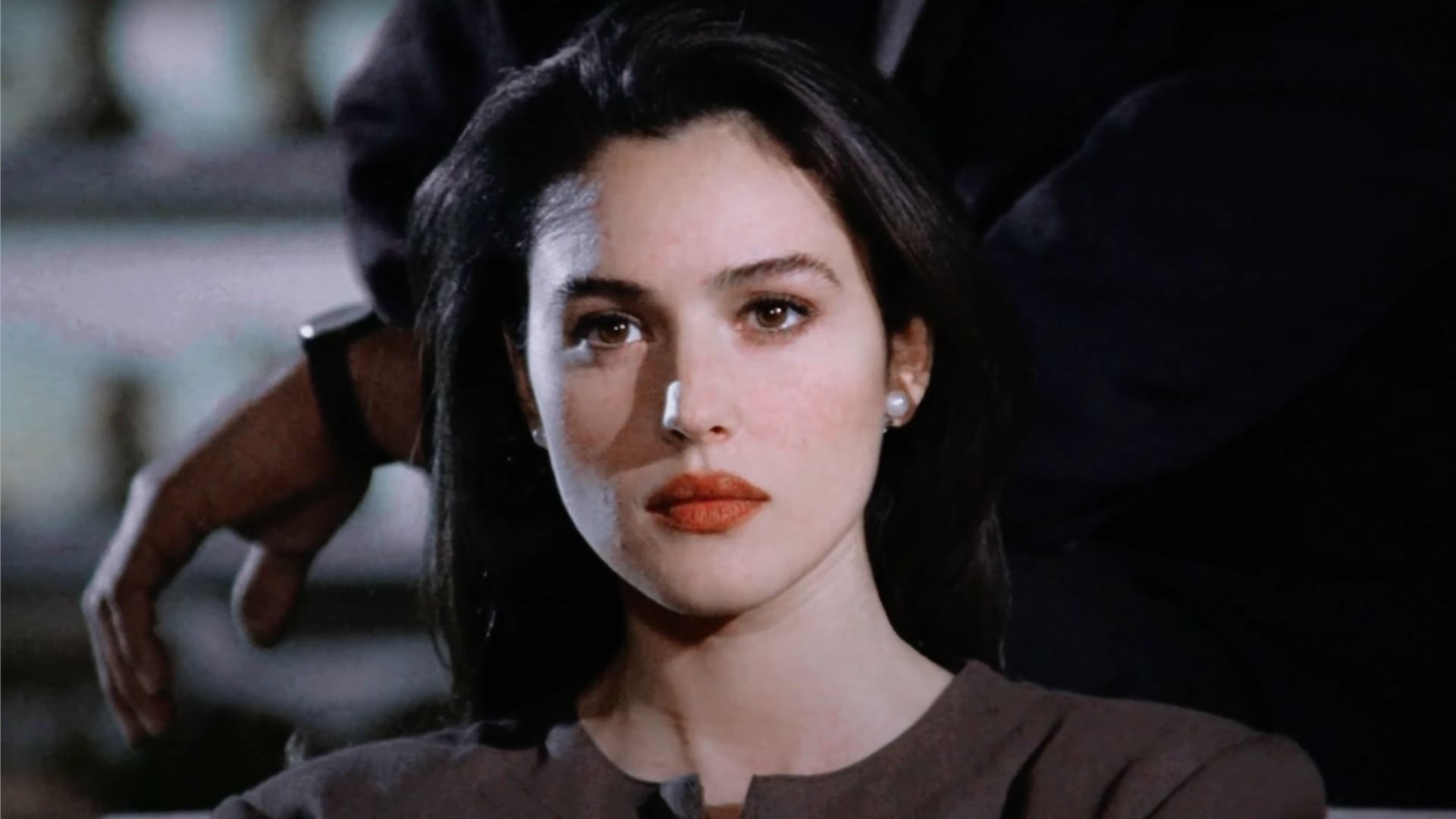 Actress Monica Bellucci with red lipstick on her lips looks to the side against the background of a man in a suit