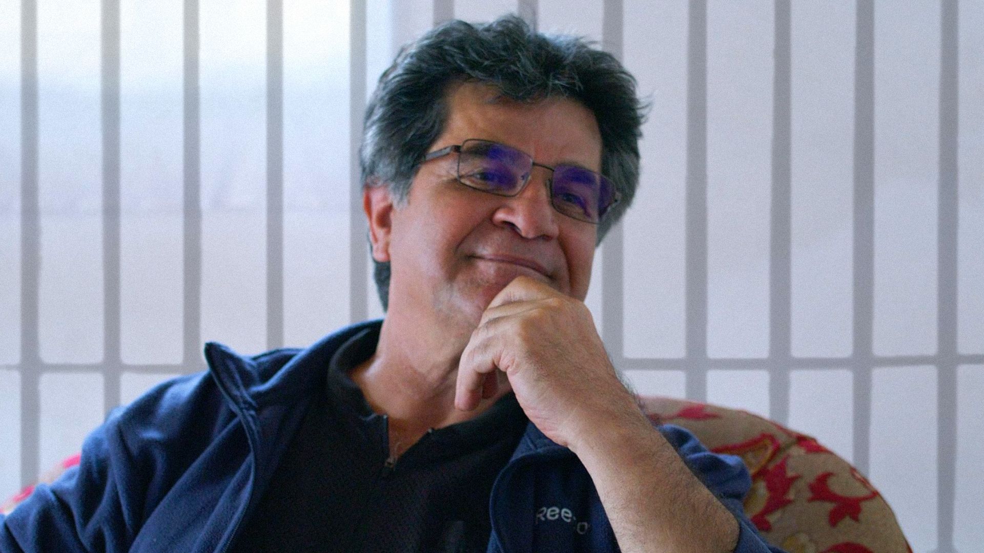 Il regista Jafar Panahi in una giacca blu è seduto su una sedia e sorride, guardando di lato