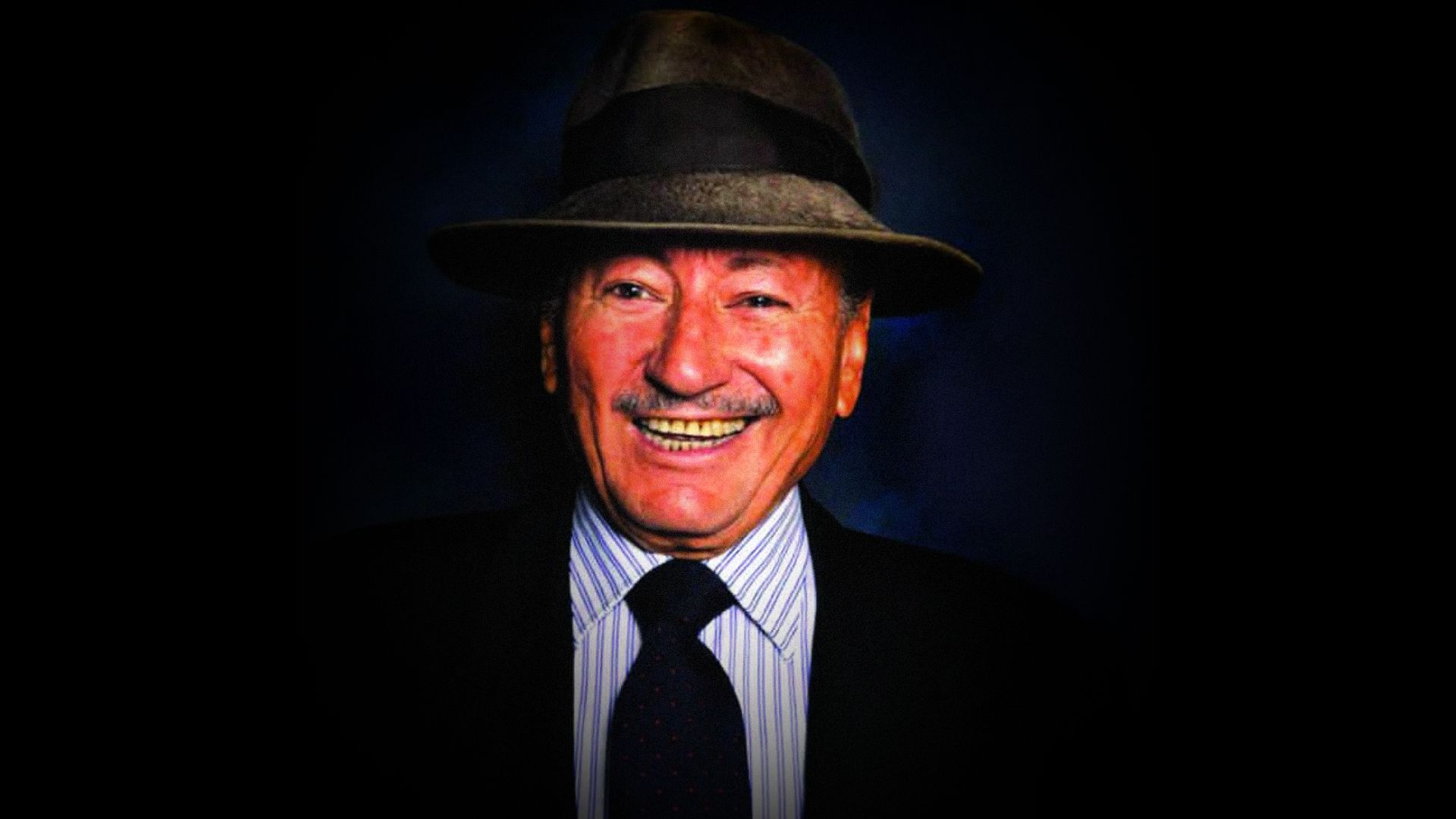 Un uomo sorridente con cappello e giacca guarda la telecamera su uno sfondo nero