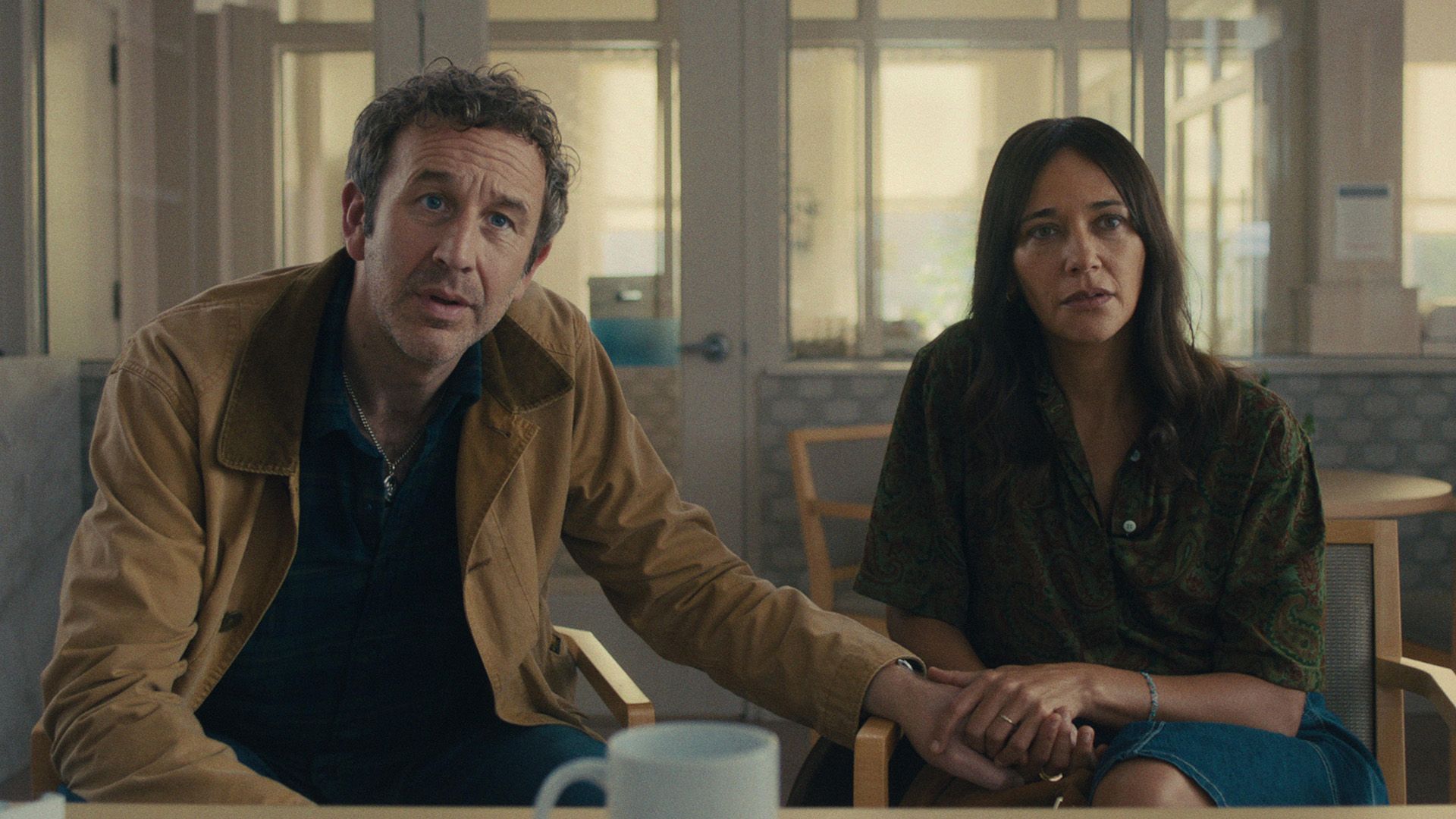 Chris O'Dowd e Rashida Jones seduti su sedie con espressioni preoccupate, tenendosi per mano