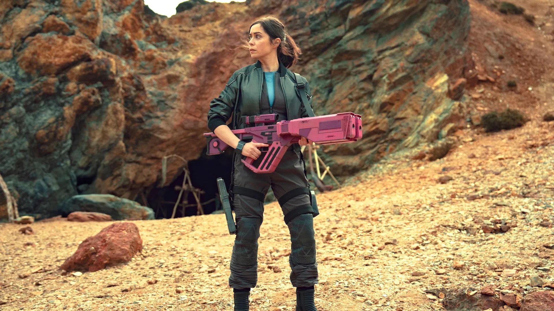 Kristin Milioti holding a pink futuristic weapon stands in a deserted landscape